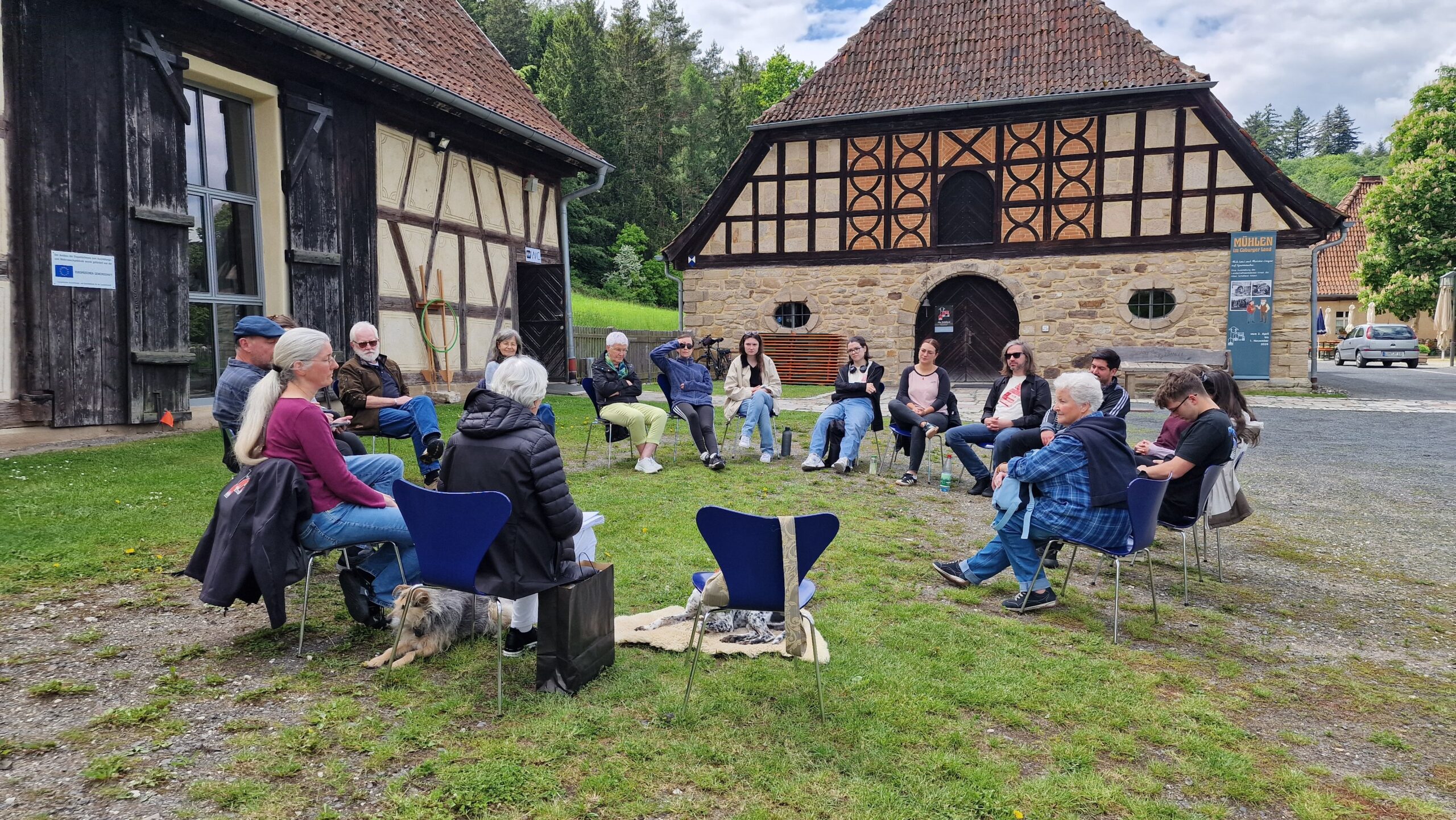 Eine Gruppe von Menschen sitzt im Freien im Kreis auf Stühlen, unterhält sich und hört zu. Sie befinden sich zwischen zwei rustikalen Gebäuden mit Fassaden aus Holz und Stein. Um sie herum erstrecken sich grünes Gras und Bäume. at Hochschule Coburg