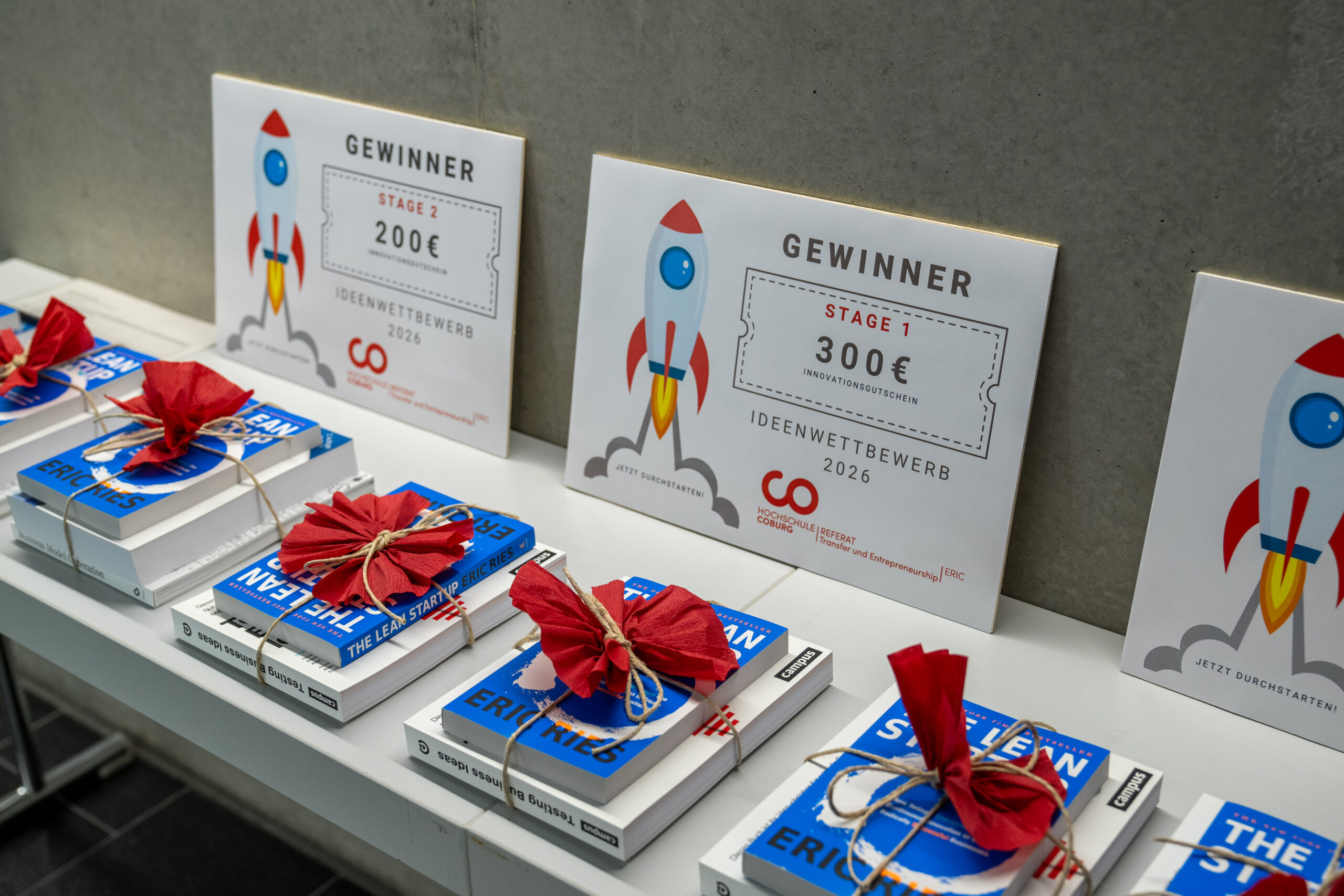 Auf einem Tisch liegen als Geschenk verpackte Bücher und Urkunden. Die Urkunden, die jeweils mit einer Raketengrafik versehen sind, weisen Geldpreise in Höhe von 200 € und 300 € für die Gewinnerin des Ideenwettbewerbs der Hochschule aus. at Hochschule Coburg