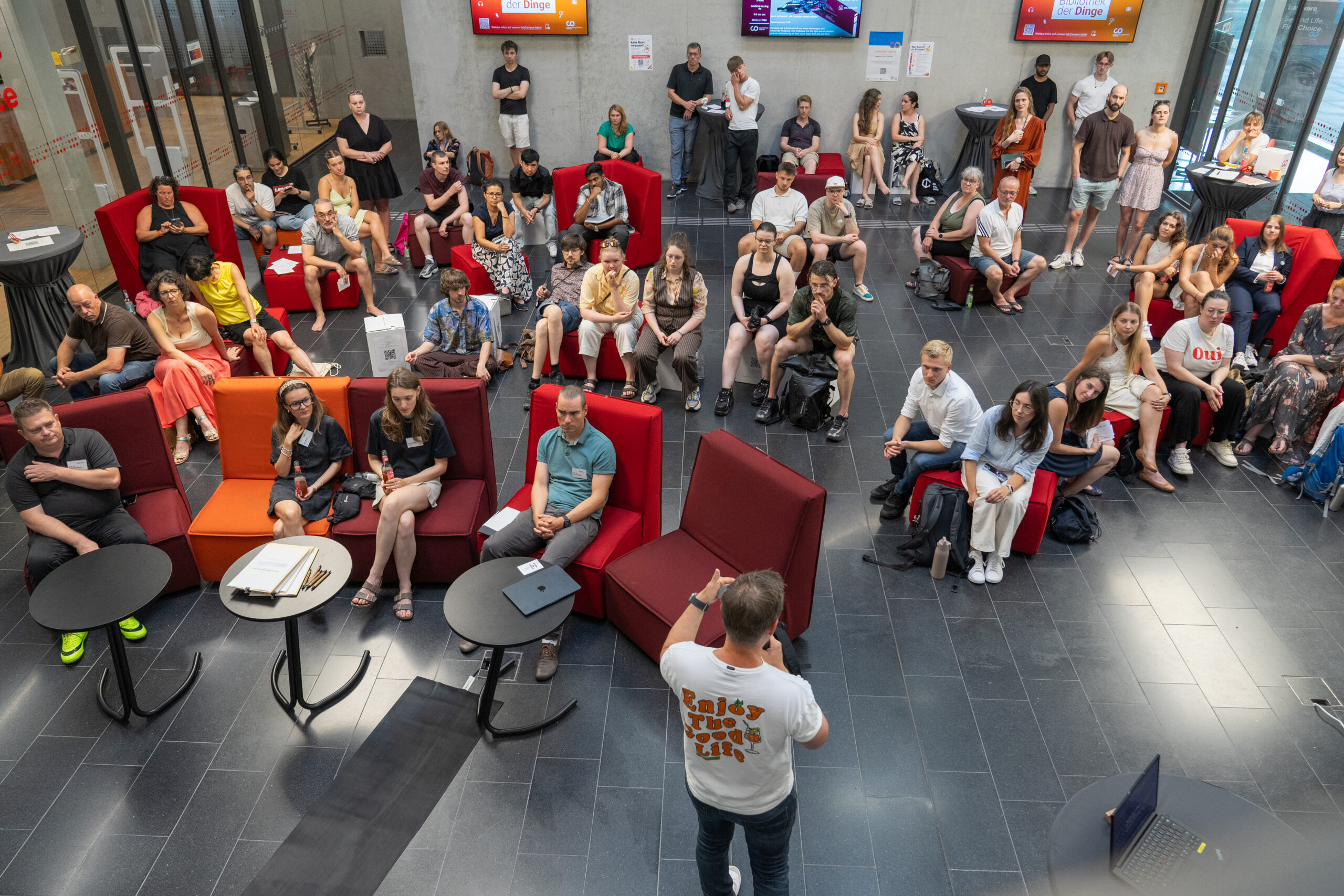 Eine Gruppe von Menschen sitzt und steht in einer modernen Lobby der Hochschule und hört einer Rednerin zu – der diesjährigen Gewinnerin des Ideenwettbewerbs –, die von vorne spricht. Einige machen sich Notizen, während andere aufmerksam zuhören. Im gesamten Raum sind leuchtend rote Stühle und Tische aufgestellt. at Hochschule Coburg