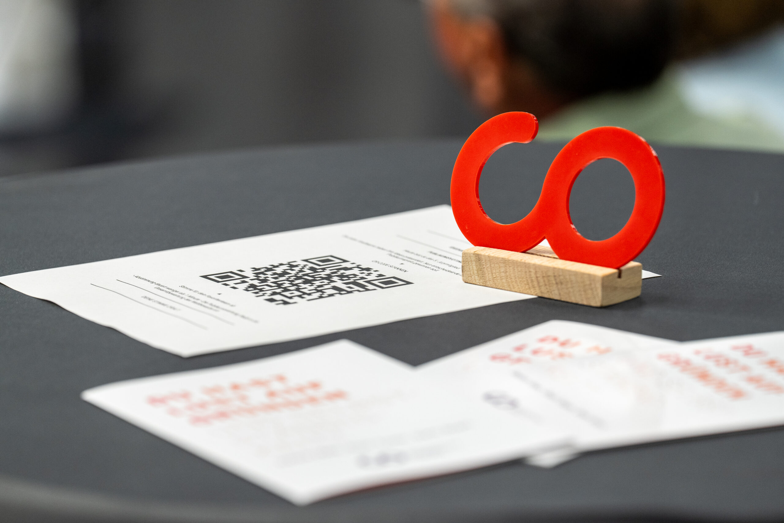 Eine rote „CO“-Skulptur, die die Gewinnerin eines Ideenwettbewerbs der Hochschule darstellt, steht auf einem Holzsockel auf einem Tisch, auf dem bedruckte Zettel und ein gut sichtbarer QR-Code verstreut liegen. Der Hintergrund ist unscharf, wodurch die Aufmerksamkeit auf die Objekte im Vordergrund gelenkt wird. at Hochschule Coburg