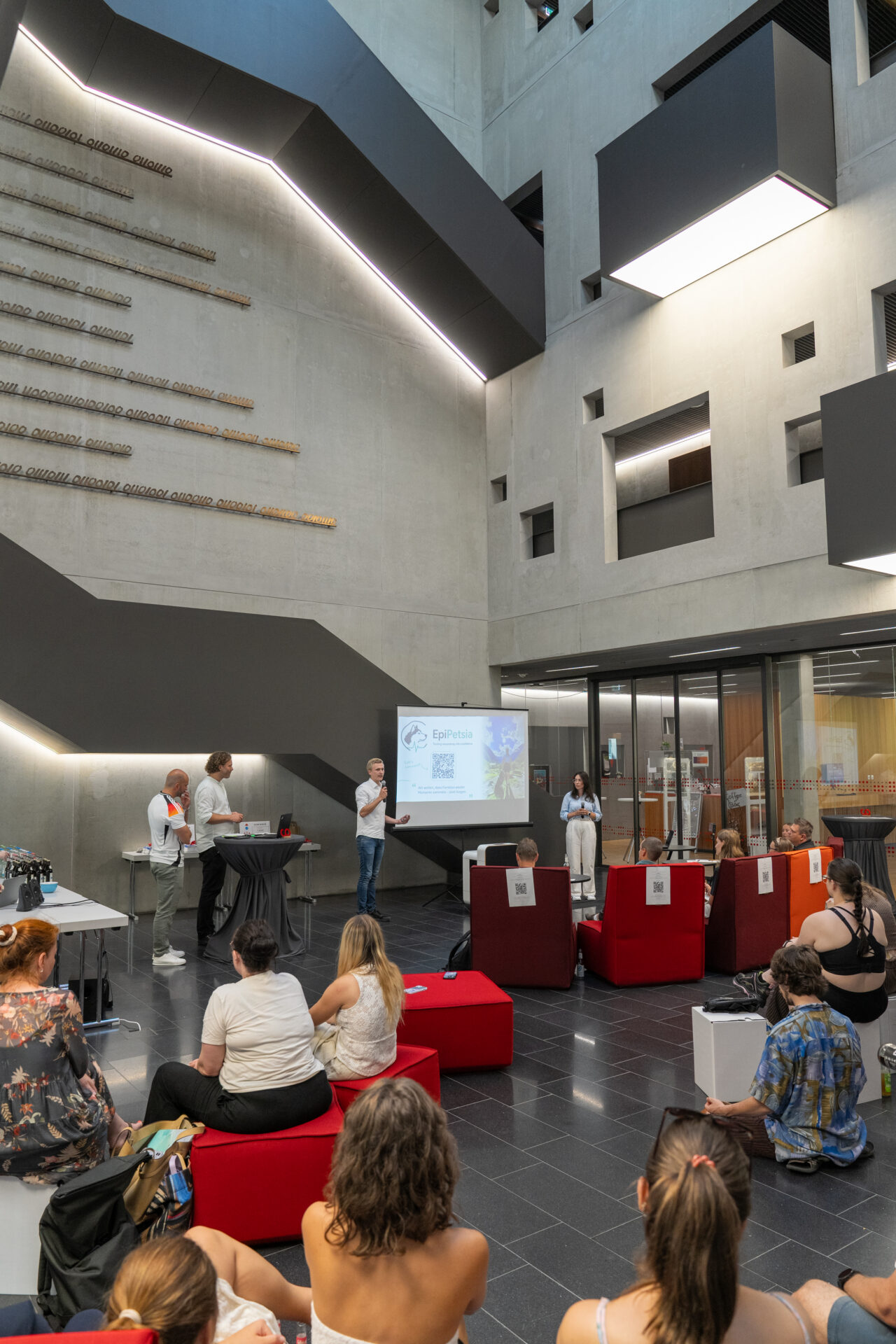 Eine Gruppe nimmt an einer Präsentation in einem modernen Atrium mit Betonwänden an der Hochschule teil. Drei Referenten stehen in der Nähe einer Projektionswand, während das Publikum, das gespannt darauf ist, mehr über die Gewinnerin des Ideenwettbewerbs zu erfahren, auf roten Bänken oder auf dem Boden sitzt und aufmerksam zuhört. at Hochschule Coburg