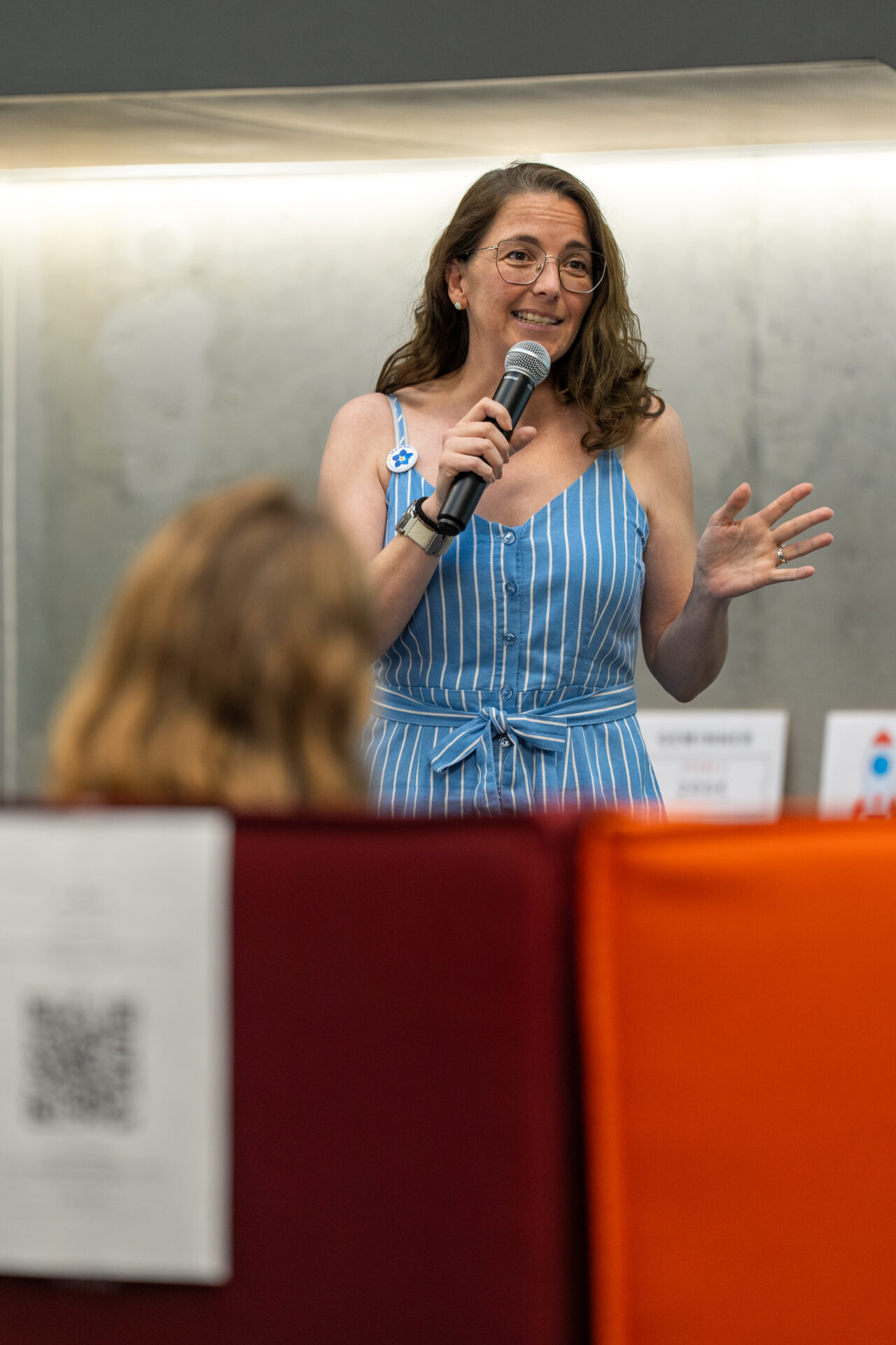Eine Frau in einem blau-weiß gestreiften Kleid spricht lächelnd und mit einer Handbewegung in ein Mikrofon, während sie als Gewinnerin des Ideenwettbewerbs in einem Raum vor einem Publikum an der Hochschule steht. at Hochschule Coburg