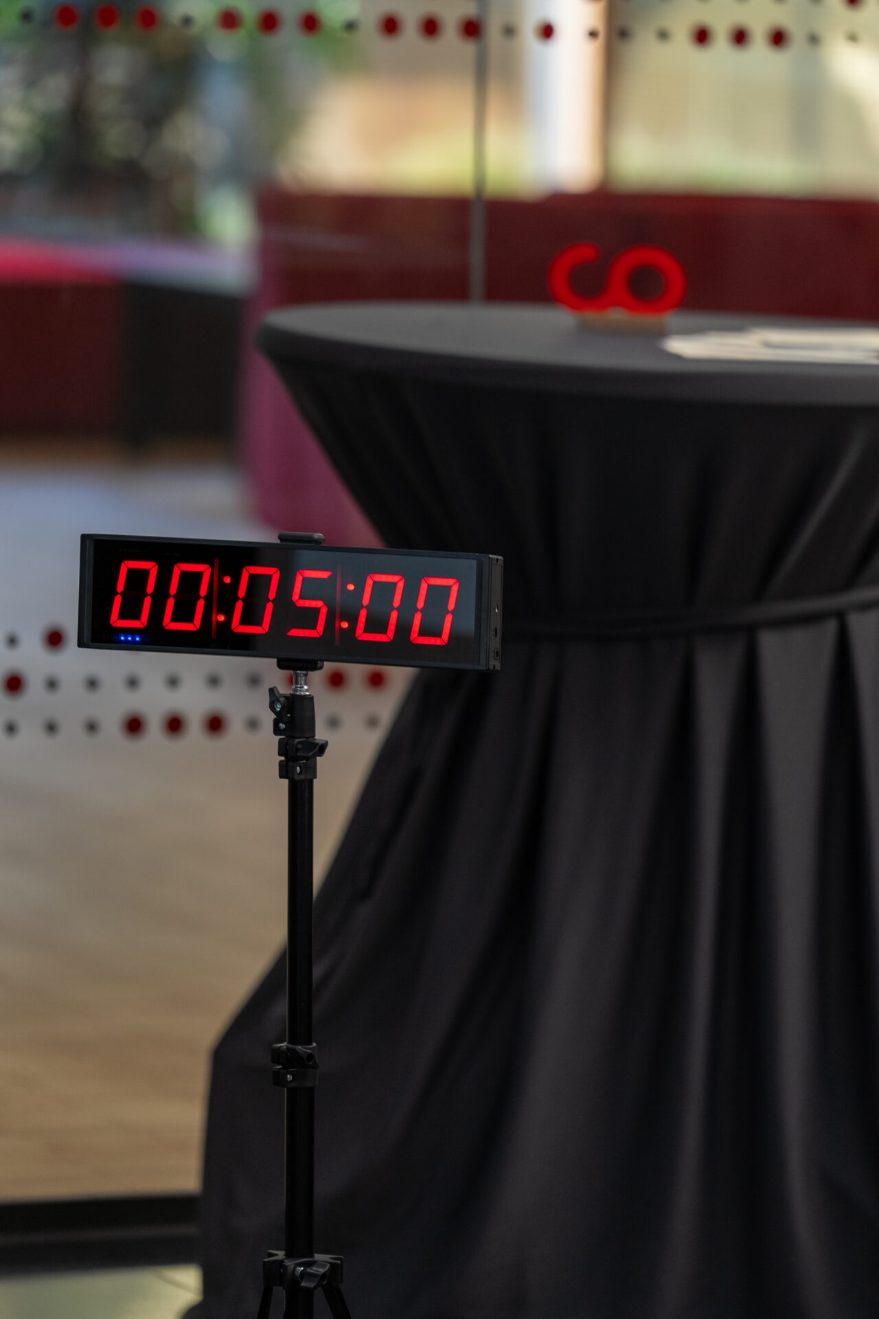 Ein digitaler Timer auf einem Ständer zeigt in roten Ziffern „00:05:00“ an, neben einem runden Tisch, der mit einem schwarzen Tischtuch bedeckt ist, in einem modernen Innenraum – eine Szene, die die Atmosphäre eines Ideenwettbewerbs an der Hochschule einfängt, bevor die Gewinnerin bekannt gegeben wird. at Hochschule Coburg