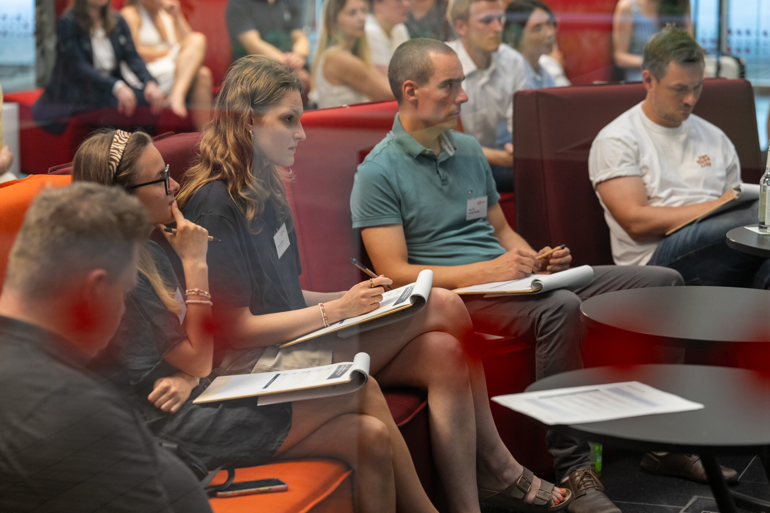 In einem modernen Raum der Hochschule sitzen Menschen auf roten Stühlen, hören aufmerksam zu und machen sich während eines Vortrags oder Workshops Notizen. Einige halten Notizblöcke und Stifte in der Hand, auf den Tischen vor ihnen liegen Unterlagen und Getränke – vielleicht inspiriert von der jüngsten Gewinnerin eines Ideenwettbewerbs. at Hochschule Coburg
