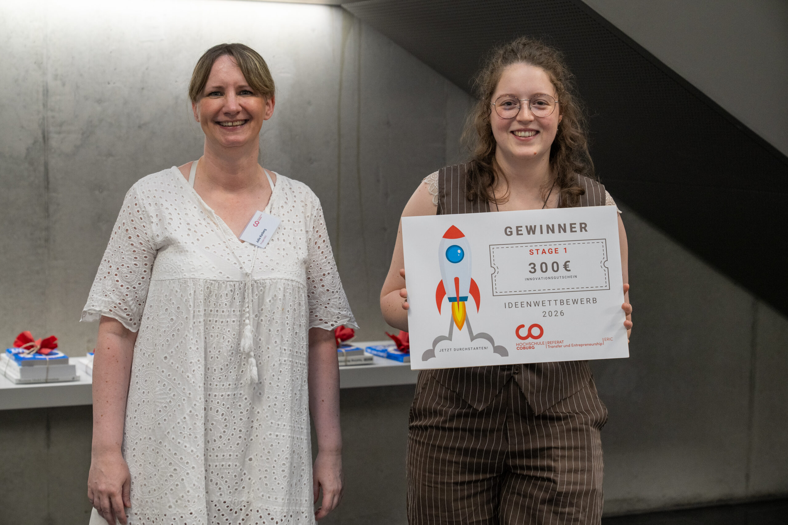 Zwei Frauen stehen in einem Raum der Hochschule; die eine lächelt, während die andere, die Gewinnerin des Ideenwettbewerbs, eine Urkunde in der Hand hält, auf der „Gewinnerin, Stufe 1, 300 €“ steht. Im Hintergrund ist vor einer Betonwand ein Tisch mit Geschenken zu sehen. at Hochschule Coburg