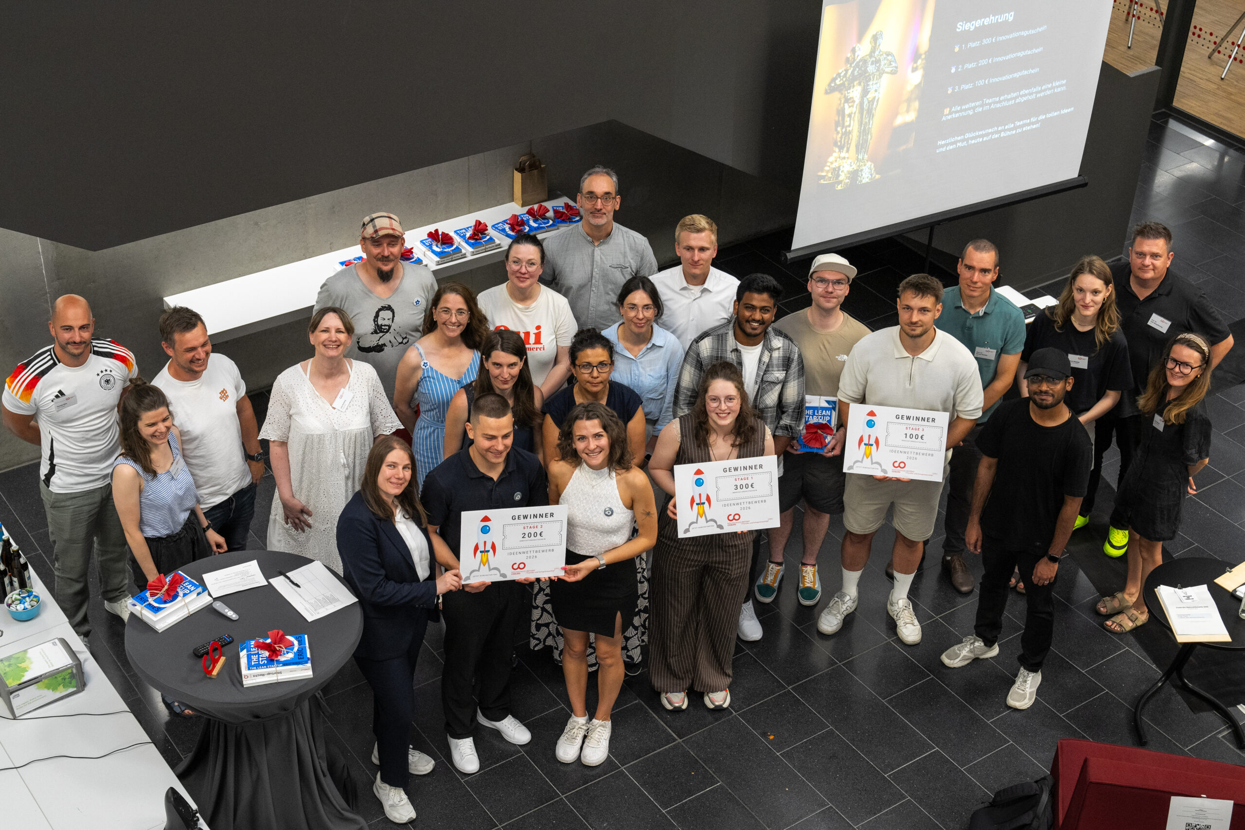 Eine Gruppe von etwa 25 Personen posiert in einem Raum, einige halten Urkunden und Pokale in den Händen. Im Hintergrund ist eine Präsentation zu sehen, auf der die Gewinnerin des Ideenwettbewerbs vorgestellt wird. Auf einem Tisch in der Nähe liegen Geschenke, und die festliche Kulisse lässt vermuten, dass diese Szene an einer Hochschule im Rahmen einer Preisverleihung aufgenommen wurde. at Hochschule Coburg