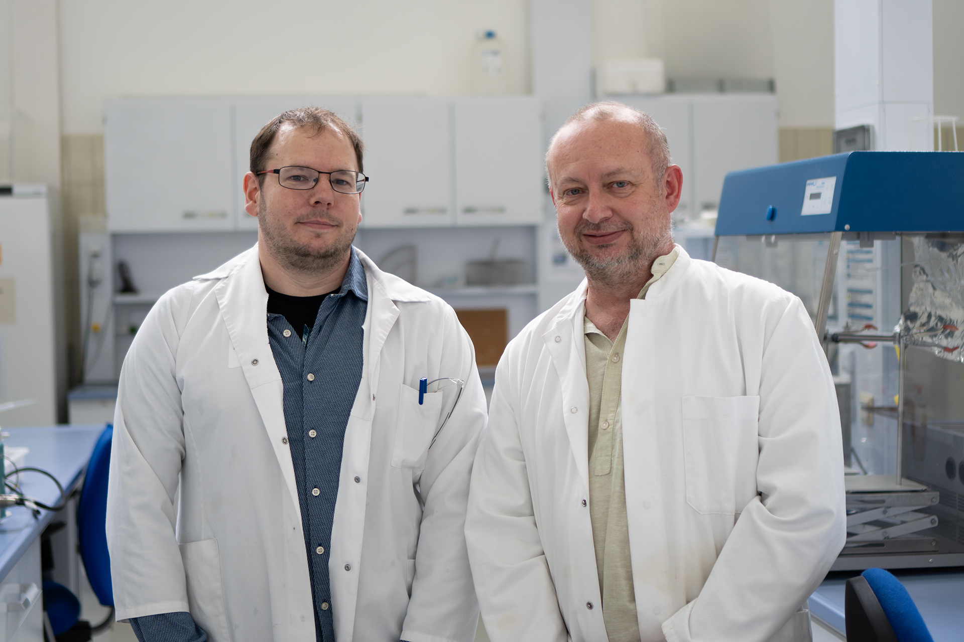 Zwei Männer in weißen Laborkitteln stehen in einem Labor, im Hintergrund sind wissenschaftliche Geräte und Schränke zu sehen. Beide blicken in die Kamera und wirken wie Forscher oder Wissenschaftler, die möglicherweise auf ihrem Karriereweg zur Professur sind und höhere akademische Positionen anstreben. at Hochschule Coburg