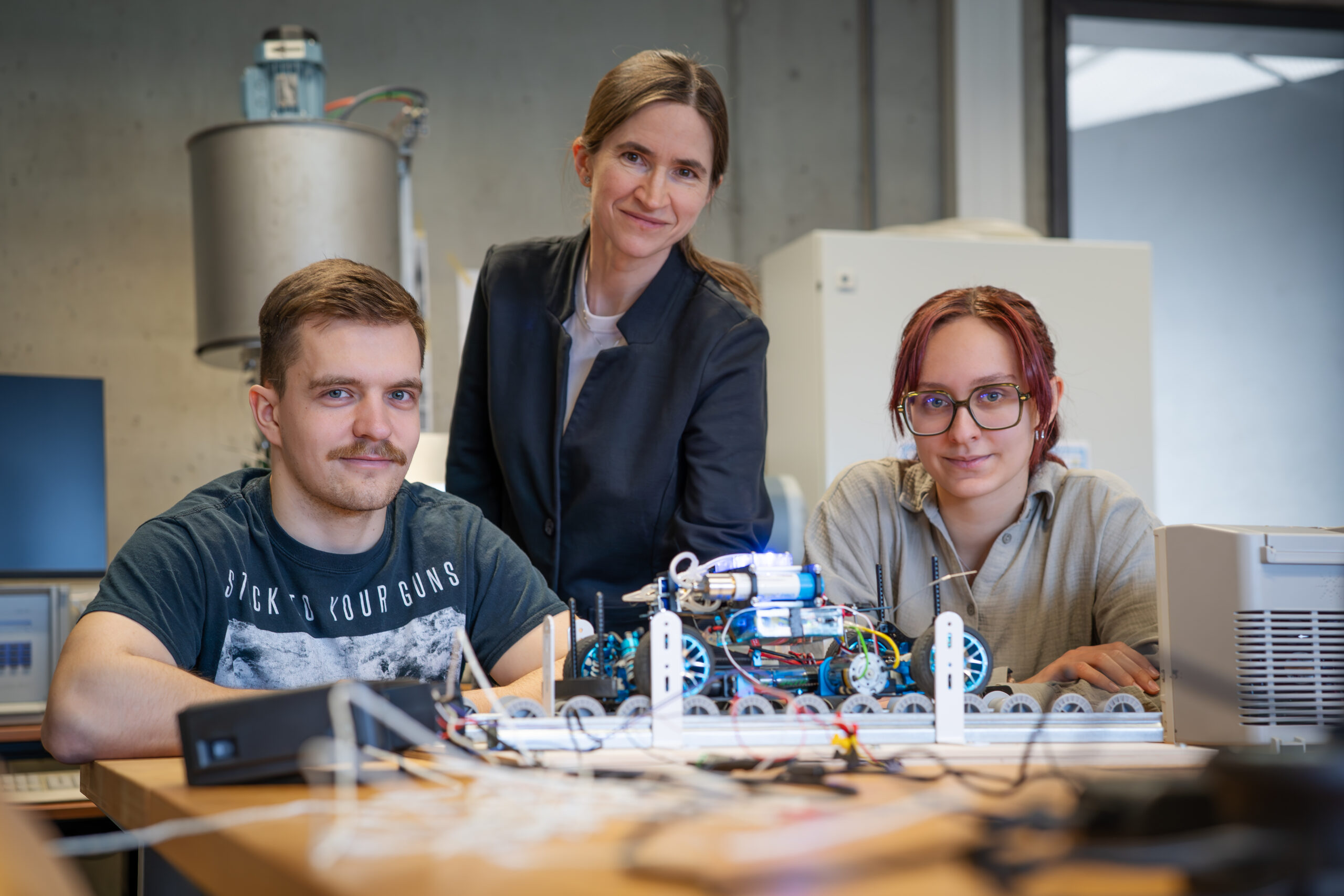 Drei Personen in einem Labor, die hinter einem Robotergerät auf einem Tisch sitzen und stehen und in die Kamera lächeln. Im Hintergrund sind Laborgeräte und Elektronik zu sehen, was eine inspirierende Umgebung für alle verdeutlicht, die eine Karriere als Professor im Bereich Robotik oder Ingenieurwesen in Betracht ziehen. at Hochschule Coburg