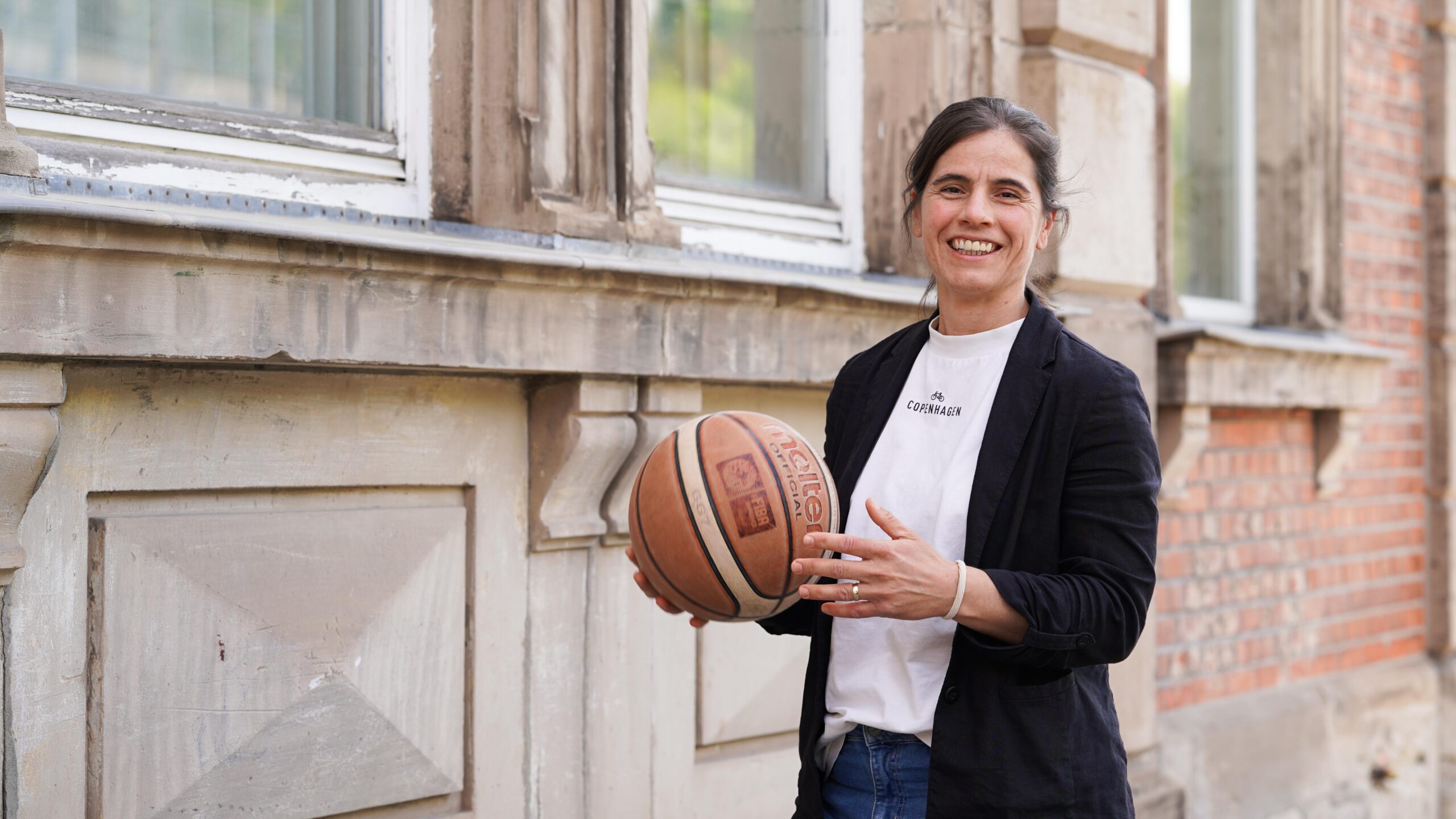 Eine lächelnde Person steht im Freien neben einem Gebäude und hält einen Basketball mit beiden Händen in den Händen. Sie trägt einen schwarzen Blazer über einem weißen Hemd und Jeans und verkörpert damit den selbstbewussten Geist der Stipendiatinnen, die voller Zuversicht auf das Jahr 2026 blicken. at Hochschule Coburg