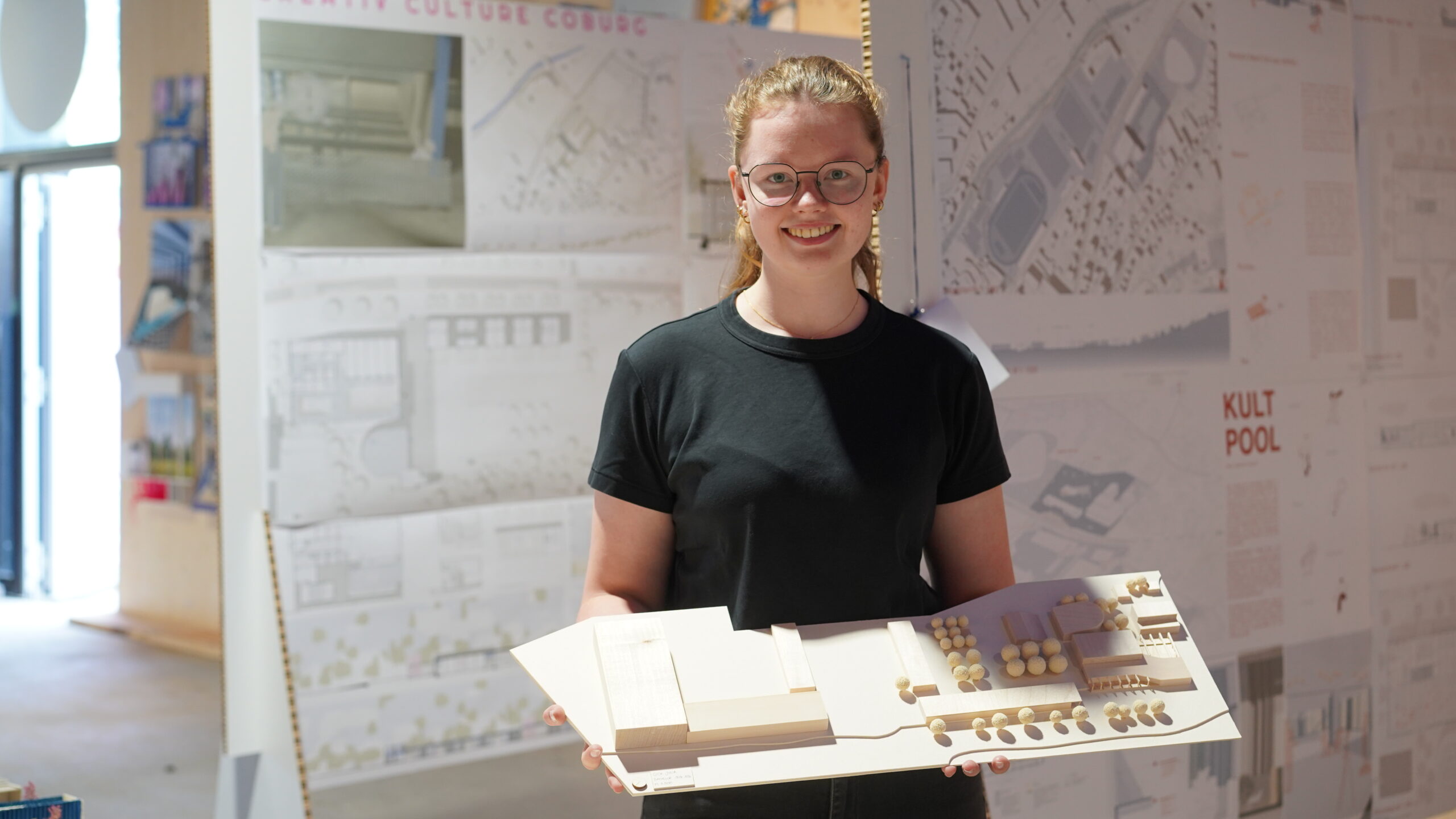 Eine Person mit Brille und schwarzem Hemd hält ein detailreiches Architekturmodell in der Hand und stellt damit ihre gestalterischen Fähigkeiten unter Beweis. Hinter ihr sind an einer Wand Architekturzeichnungen und Plakate zu sehen, die die kreative Atmosphäre einer Werkschau-Ausstellung widerspiegeln. at Hochschule Coburg
