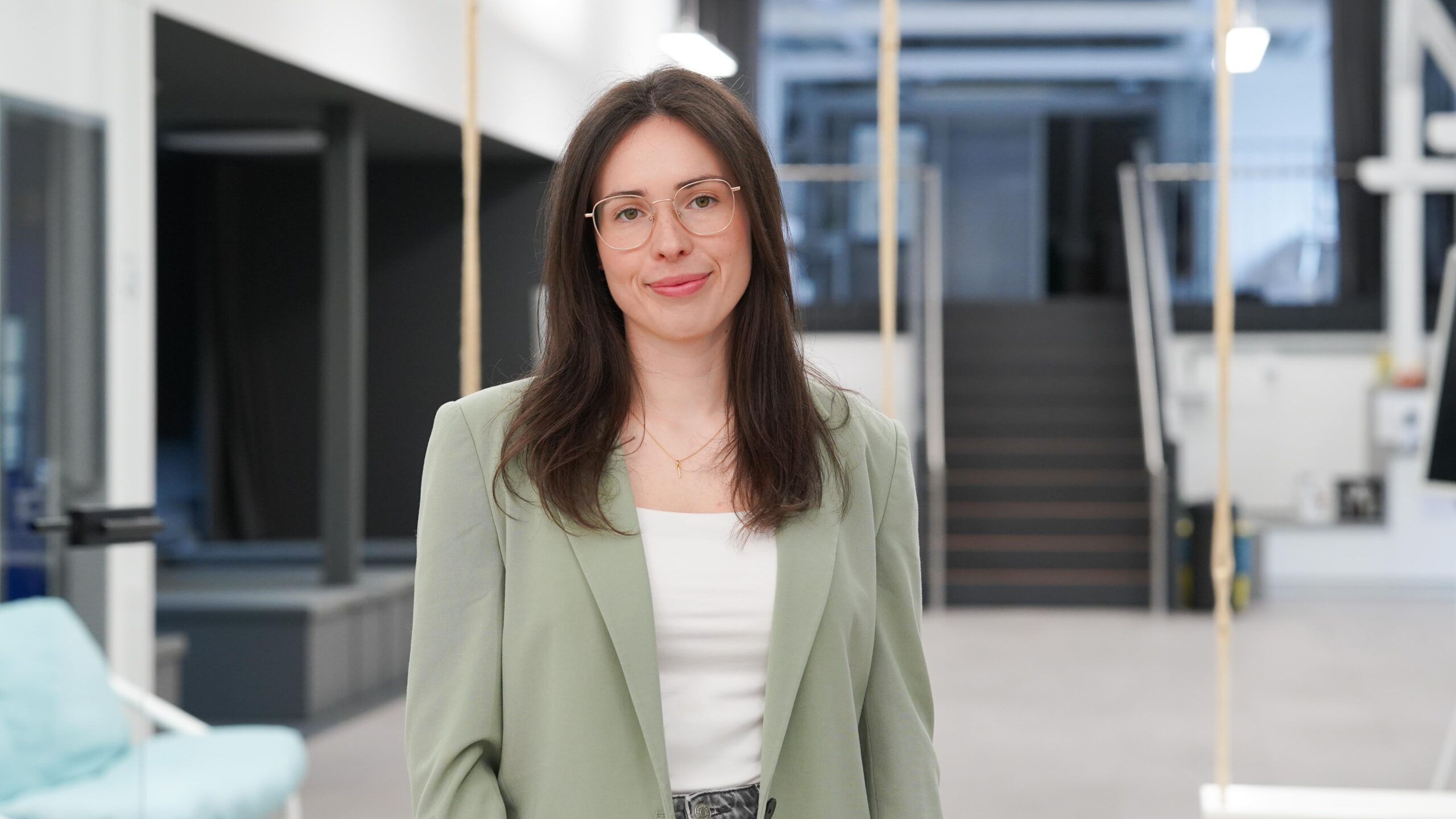 Eine Frau mit langen braunen Haaren und Brille, die einen hellgrünen Blazer über einem weißen Oberteil trägt, steht in einem modernen, hellen Büroraum, im Hintergrund sind Treppen zu sehen – eine inspirierende Szene, die die dynamischen Stipendiatinnen des Jahres 2026 gut verkörpern könnte. at Hochschule Coburg
