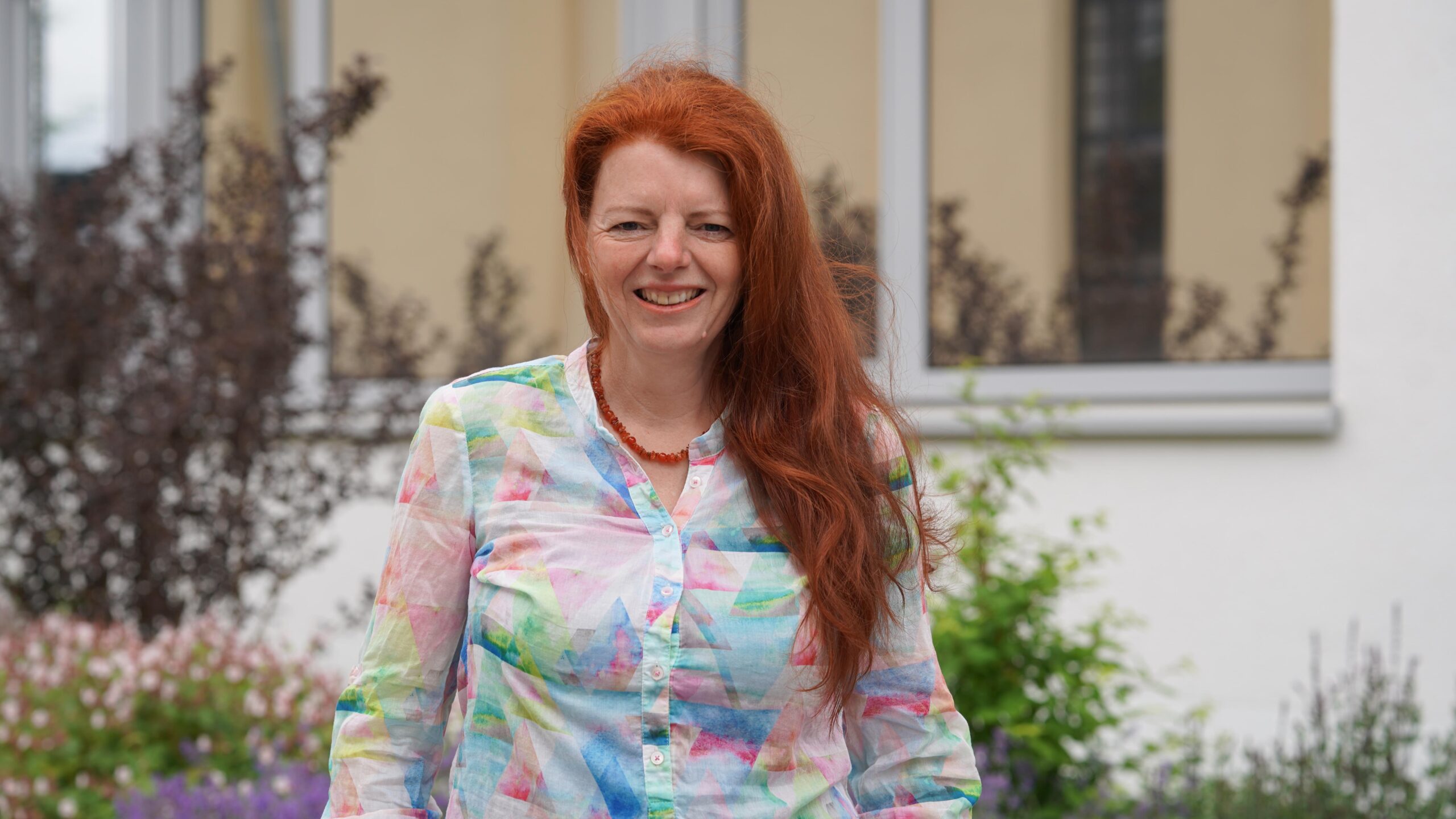 Eine Frau mit langen roten Haaren, die ein buntes Hemd mit geometrischen Mustern trägt, lächelt im Freien vor einem Gebäude mit Fenstern und Gartenpflanzen – sie ist eine der Stipendiatinnen des Jahres 2026. at Hochschule Coburg