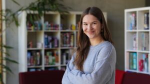 Eine junge Frau mit langen braunen Haaren, die einen hellgrauen Pullover trägt, lächelt mit vor der Brust verschränkten Armen vor einem Bücherregal voller Bücher und Spiele – eine selbstbewusste Stipendiatin des Jahrgangs 2026, die ihr akademisches Umfeld genießt. at Hochschule Coburg