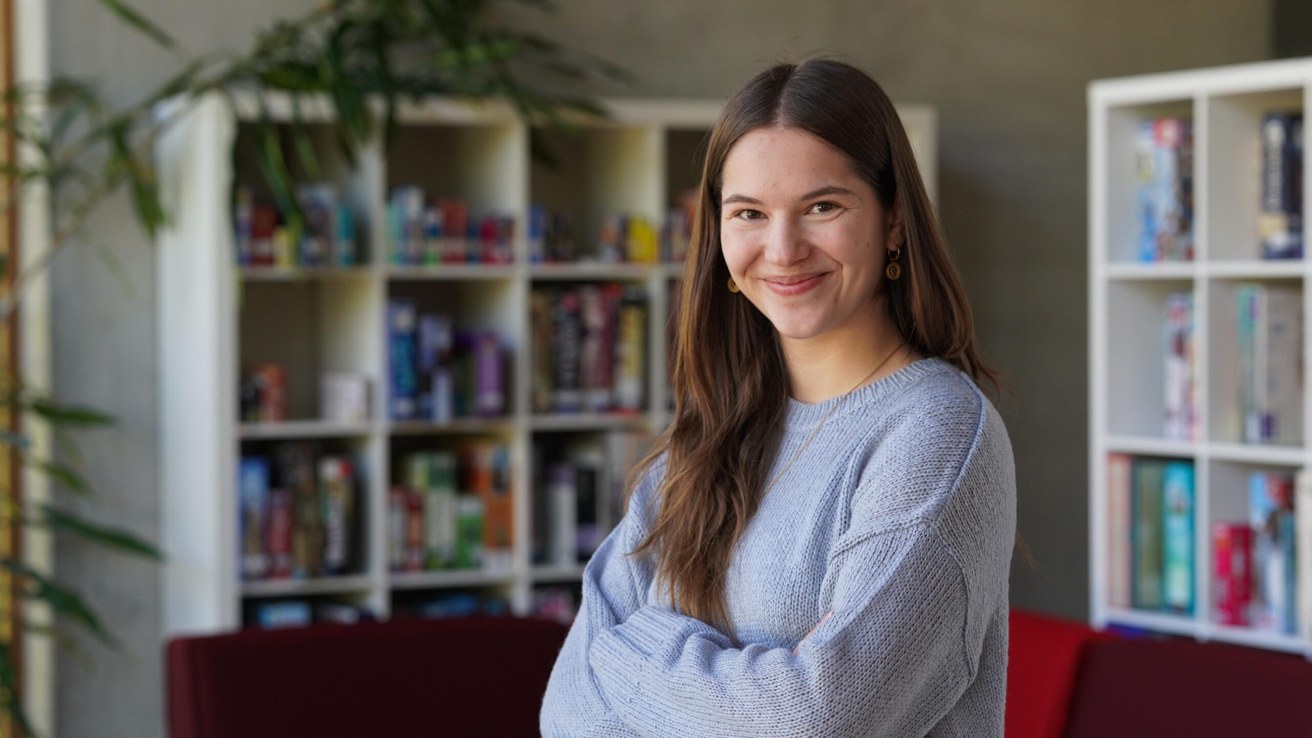 Eine junge Frau mit langen braunen Haaren, die einen hellgrauen Pullover trägt, lächelt mit vor der Brust verschränkten Armen vor einem Bücherregal voller Bücher und Spiele – eine selbstbewusste Stipendiatin des Jahrgangs 2026, die ihr akademisches Umfeld genießt. at Hochschule Coburg
