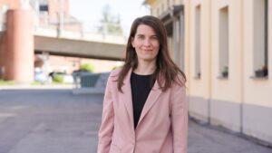 Eine Frau mit langen braunen Haaren, die einen hellrosa Blazer und ein schwarzes Oberteil trägt, steht draußen in der Nähe eines beigen Gebäudes und lächelt in die Kamera. Als eine der Stipendiatinnen des Jahres 2026 wirkt sie selbstbewusst und gelassen. Im Hintergrund sind eine Straße, eine Brücke und etwas Grün zu sehen. at Hochschule Coburg