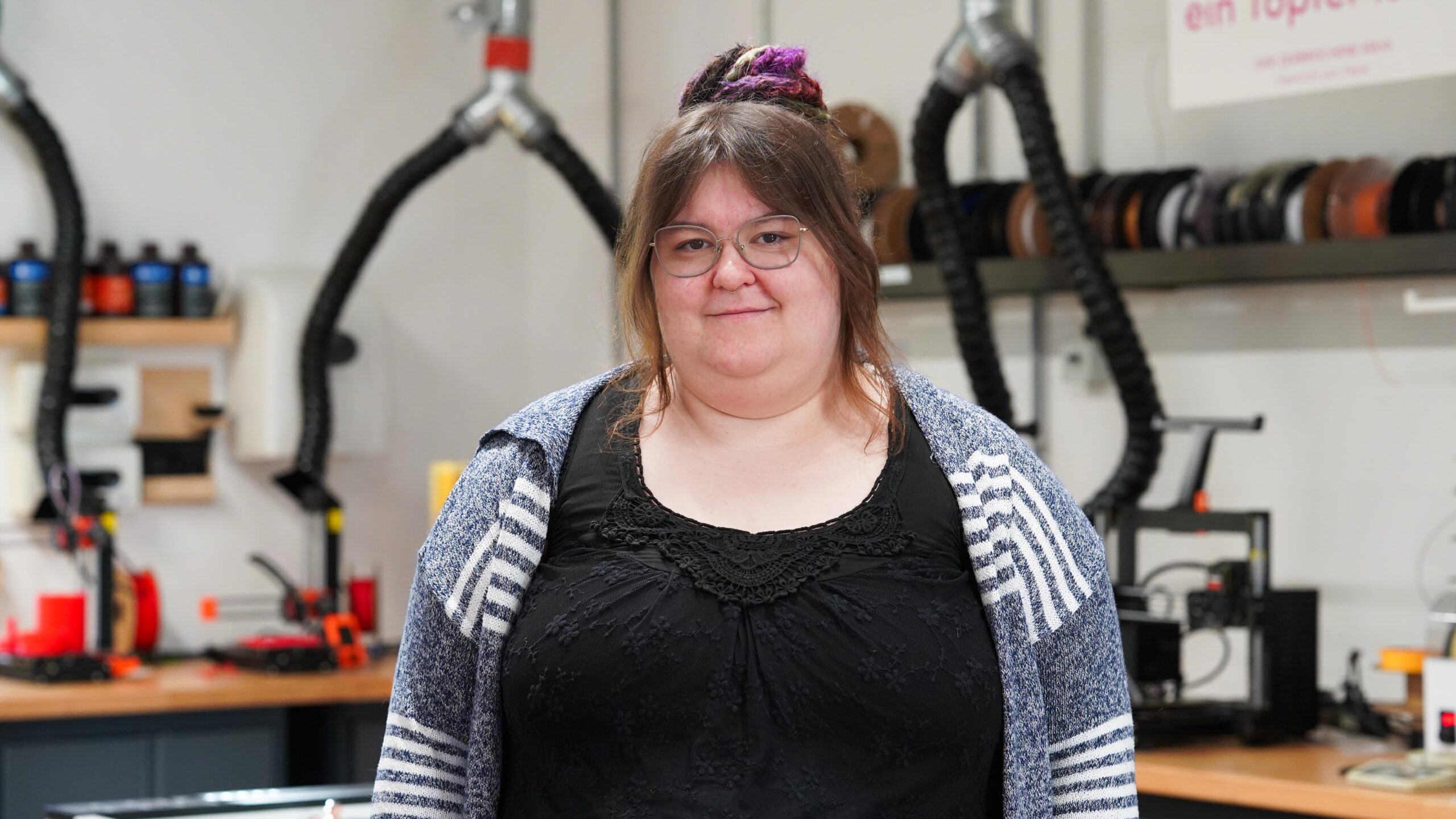 Eine Person mit Brille und langen braunen Haaren, die ein schwarzes Oberteil und einen gestreiften Pullover trägt, steht in einer Werkstatt, im Hintergrund sind Werkzeuge, Regale und Geräte zu sehen – als Symbol für den Innovationsgeist der Stipendiatinnen 2026. at Hochschule Coburg