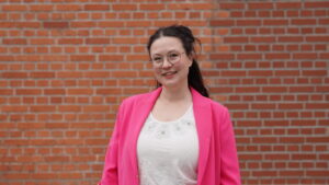 Eine Frau mit langen dunklen Haaren, Brille und einem leuchtend rosa Blazer über einem weißen Oberteil steht lächelnd vor einer roten Backsteinmauer und verkörpert das Selbstbewusstsein und den Ehrgeiz der Stipendiatinnen des Jahrgangs 2026. at Hochschule Coburg