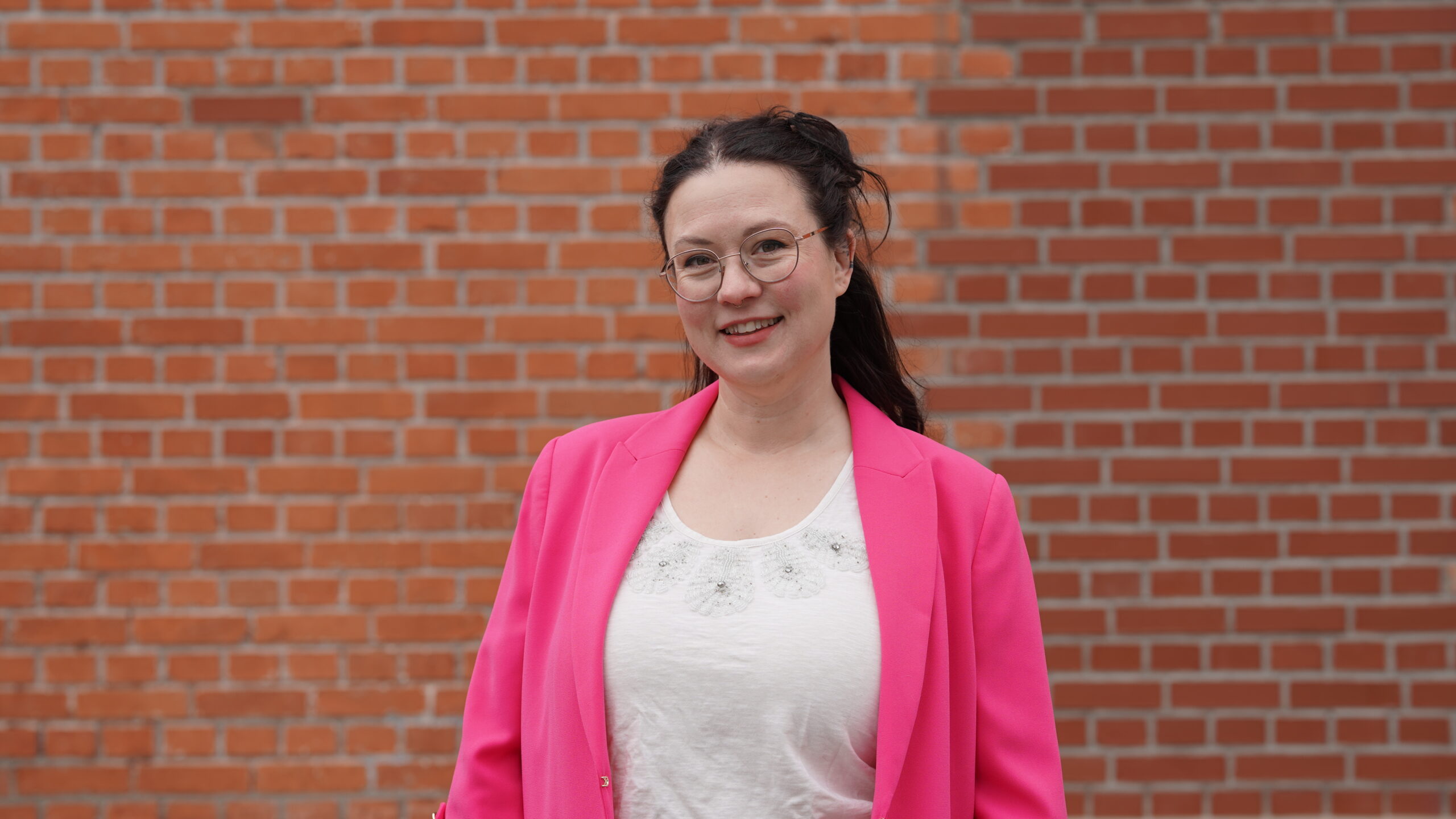 Eine Frau mit langen dunklen Haaren, Brille und einem leuchtend rosa Blazer über einem weißen Oberteil steht lächelnd vor einer roten Backsteinmauer und verkörpert das Selbstbewusstsein und den Ehrgeiz der Stipendiatinnen des Jahrgangs 2026. at Hochschule Coburg