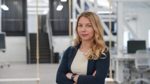 Eine Frau mit langen blonden Haaren, die einen dunklen Blazer und ein helles Oberteil trägt, steht mit verschränkten Armen in einem modernen Büroraum – mit Schreibtischen, Computern und einer Treppe im Hintergrund –, der das berufliche Umfeld von „Stipendiatinnen 2026“ widerspiegelt. at Hochschule Coburg