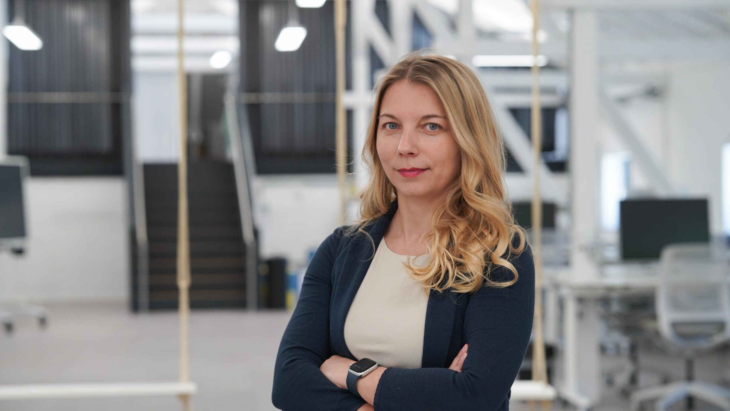 Eine Frau mit langen blonden Haaren, die einen dunklen Blazer und ein helles Oberteil trägt, steht mit verschränkten Armen in einem modernen Büroraum – mit Schreibtischen, Computern und einer Treppe im Hintergrund –, der das berufliche Umfeld von „Stipendiatinnen 2026“ widerspiegelt. at Hochschule Coburg