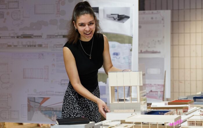 Eine Frau lächelt, während sie ein Architekturmodell präsentiert, umgeben von anderen maßstabsgetreuen Modellen und Bauplänen in einer Atelierkulisse im Rahmen der Werkschau der Fakultät Design. at Hochschule Coburg