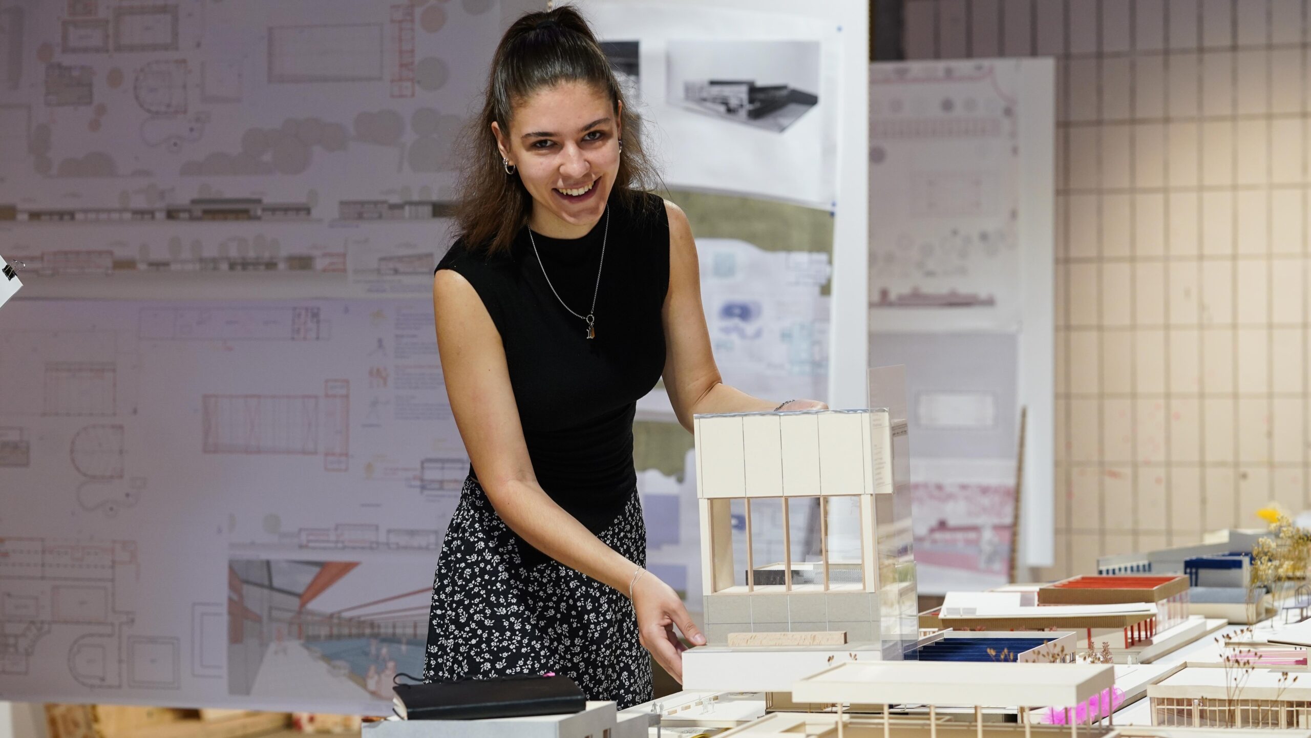 Eine Frau lächelt, während sie ein Architekturmodell präsentiert, umgeben von anderen maßstabsgetreuen Modellen und Bauplänen in einer Atelierkulisse im Rahmen der Werkschau der Fakultät Design. at Hochschule Coburg