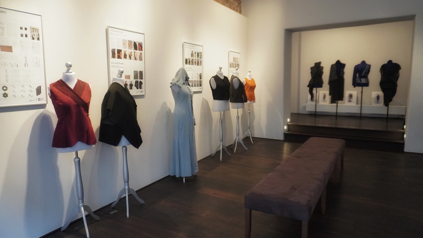 Eine Modeausstellung im Rahmen der WERKSCHAU 2026 zeigt verschiedene Kleidungsstücke auf Schaufensterpuppen vor weißen Wänden, darüber hängen gerahmte Entwurfsskizzen. In der Mitte steht eine Bank, die die Besucher dazu einlädt, während der Vernissage eine Pause einzulegen und in Gedanken zu versinken. Im hinteren Bereich werden in einer Nische weitere Outfits unter dem Motto „FASHION OPEN`N CLOSED“ präsentiert. at Hochschule Coburg