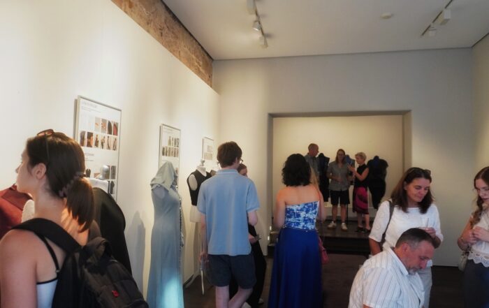 In einem hell beleuchteten Galerieraum mit hellen Wänden betrachten die Besucher die Ausstellungsstücke und unterhalten sich, während Kunst- oder Modekreationen von FASHION OPEN`N CLOSED sorgfältig auf Schaufensterpuppen arrangiert sind. Im Hintergrund hat sich eine Gruppe in der Nähe eines Backsteinbogens versammelt und fängt so die lebhafte Atmosphäre dieser Vernissage auf der WERKSCHAU 2026 ein. at Hochschule Coburg