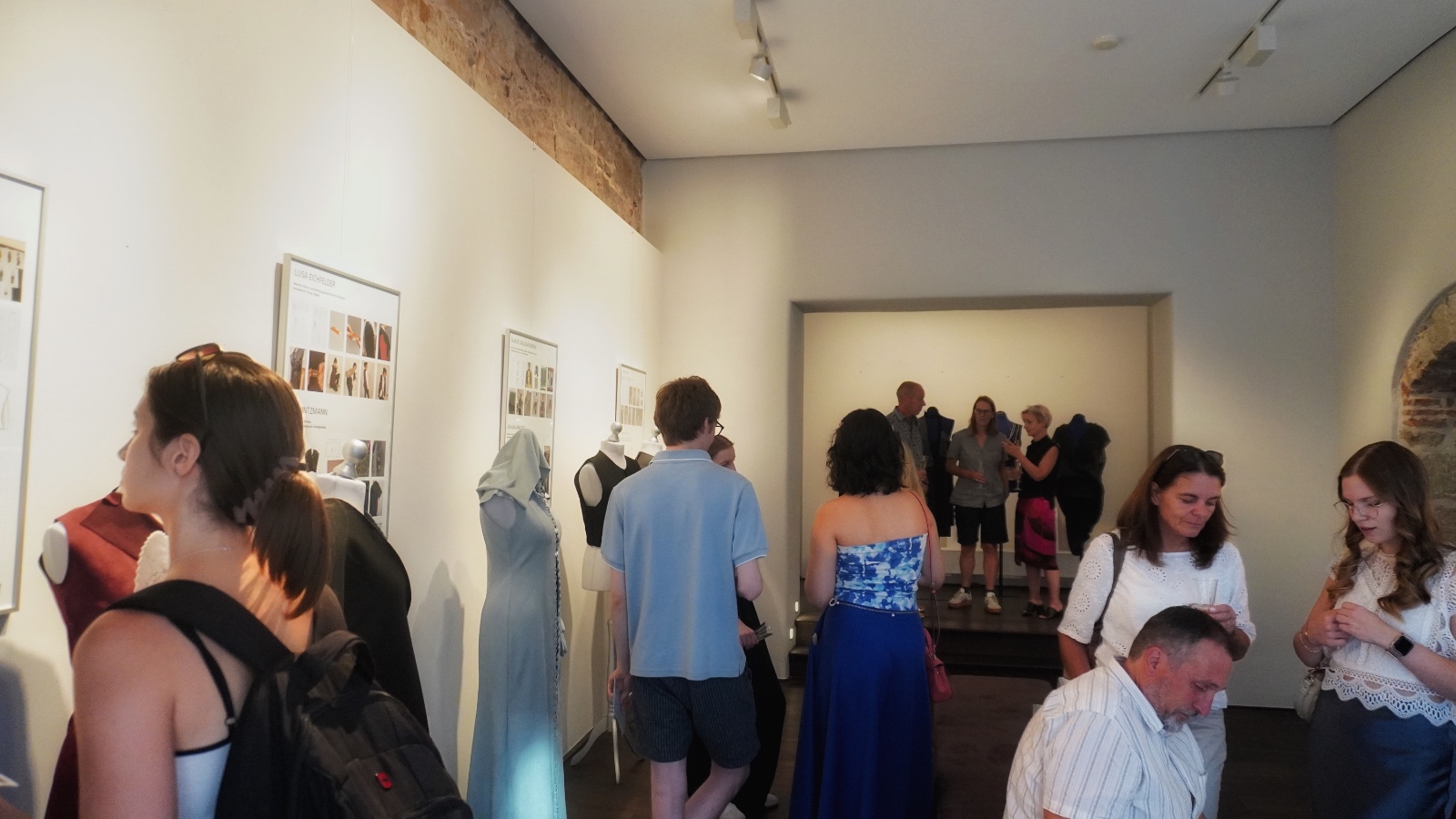 In einem hell beleuchteten Galerieraum mit hellen Wänden betrachten die Besucher die Ausstellungsstücke und unterhalten sich, während Kunst- oder Modekreationen von FASHION OPEN`N CLOSED sorgfältig auf Schaufensterpuppen arrangiert sind. Im Hintergrund hat sich eine Gruppe in der Nähe eines Backsteinbogens versammelt und fängt so die lebhafte Atmosphäre dieser Vernissage auf der WERKSCHAU 2026 ein. at Hochschule Coburg