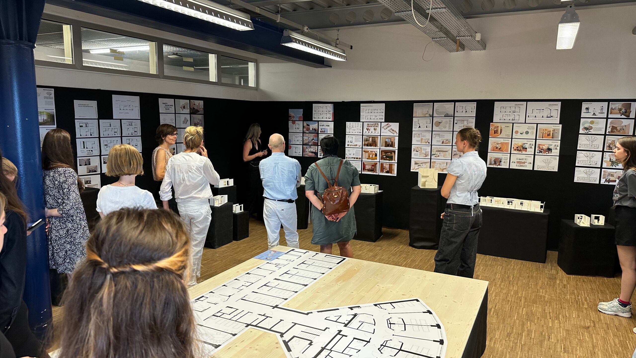 Eine Gruppe von Menschen steht in einem Raum und betrachtet während der Ausstellung der Fakultät Design + Bauen Architekturzeichnungen und Modelle, die an Wänden und auf Tischen ausgestellt sind, und beschäftigt sich mit den auf der Werkschau präsentierten kreativen Arbeiten. at Hochschule Coburg