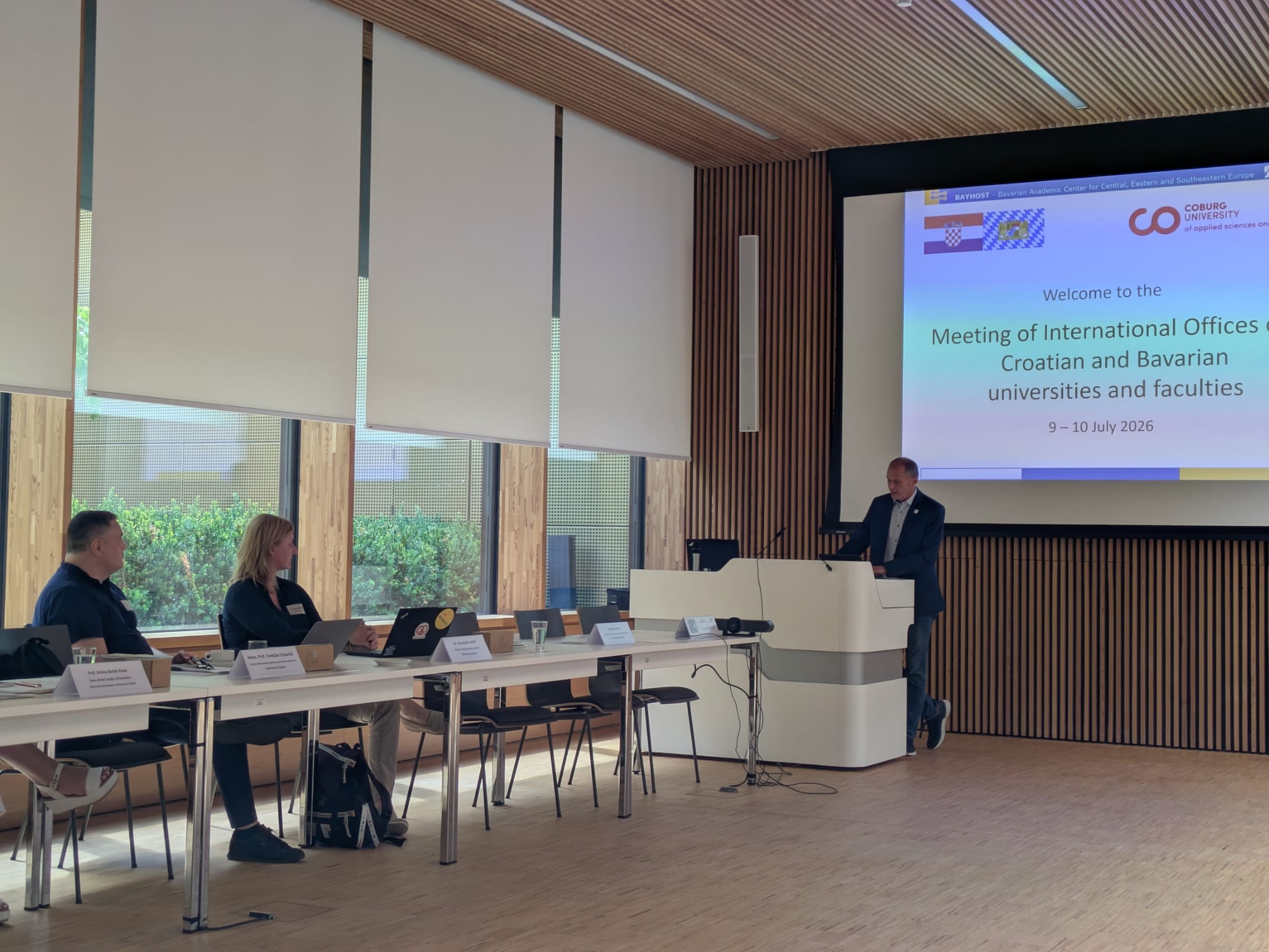 Bei einem Treffen von Vertretern kroatischer und bayerischer Hochschulen, das von deren Internationalen Büros organisiert wurde, spricht ein Mann vom Rednerpult aus zu zwei sitzenden Teilnehmern, während in einem modernen Konferenzraum eine Präsentation auf einer Leinwand gezeigt wird. at Hochschule Coburg