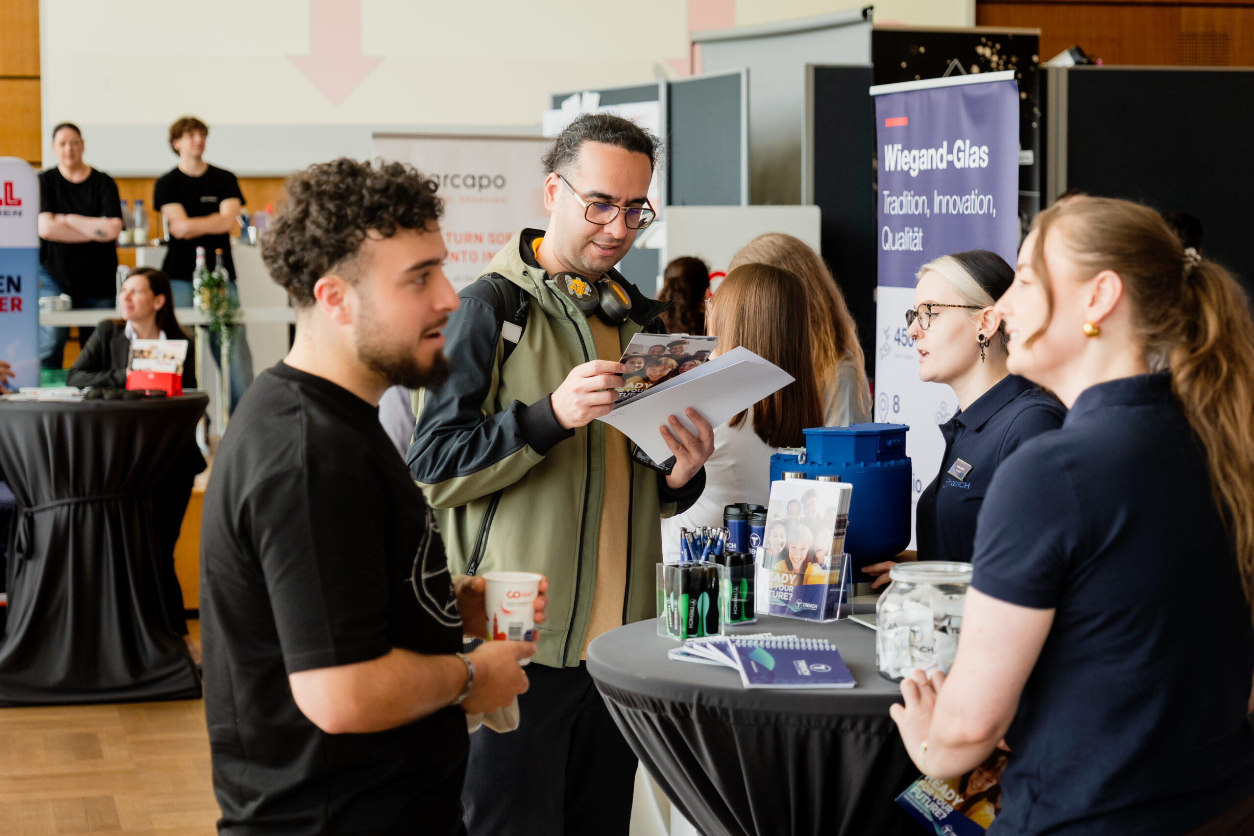 An einem Messestand tauschen sich Menschen aus; dort sind Broschüren, Werbeartikel und Banner ausgestellt. Ein Mann schaut sich eine Broschüre an, während er sich mit zwei Frauen hinter einem Tisch unterhält. Die Atmosphäre ist lebhaft und professionell. at Hochschule Coburg