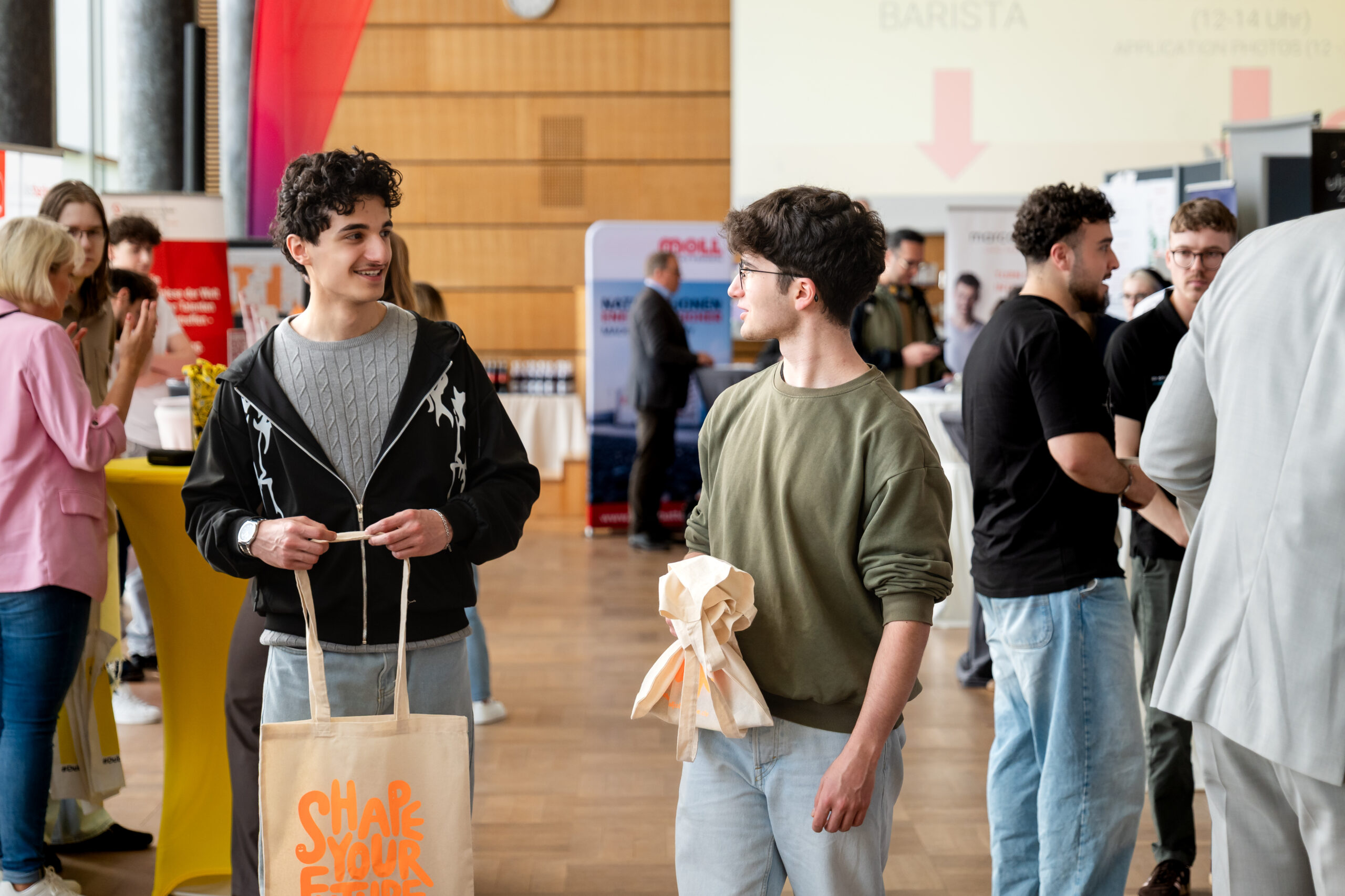 Zwei junge Männer stehen bei einer gut besuchten Veranstaltung in einem Saal, halten Tragetaschen in den Händen und unterhalten sich. Im Hintergrund mischen sich die Besucher unter den Tischen und Informationsständen, die in helles Licht getaucht sind. at Hochschule Coburg