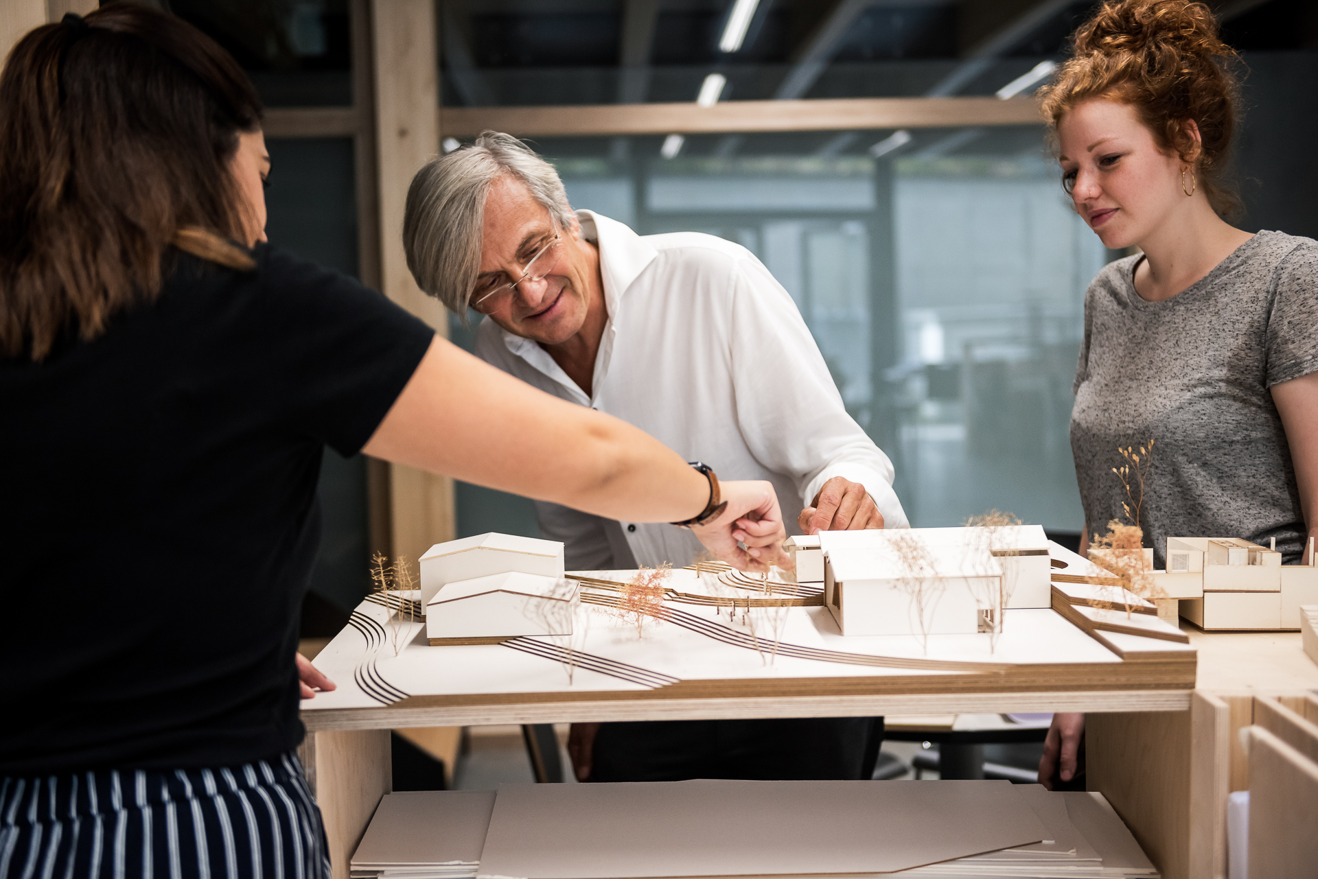 Drei Personen stehen um ein Architekturmodell herum und besprechen Gestaltungsdetails. Der Mann in der Mitte lächelt, während er auf das Modell zeigt; die beiden Frauen beobachten das Geschehen, beteiligen sich an der Diskussion und denken darüber nach, wie solche Gemeinschaftsprojekte einen Karriereweg als Professor prägen können. at Hochschule Coburg