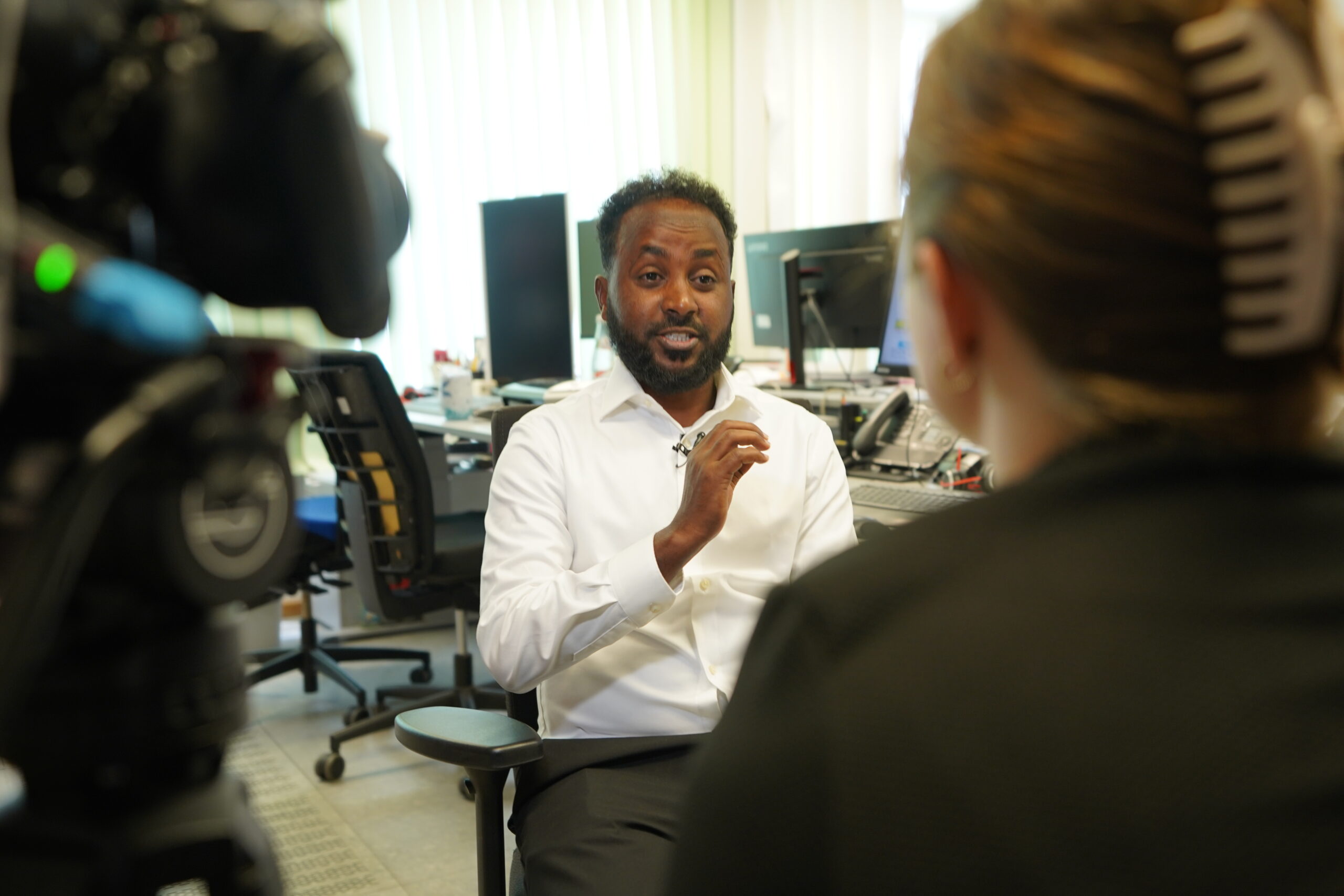 Ein Mann in einem weißen Hemd spricht während eines Interviews in einem Büro und blickt dabei auf eine Frau, die der Kamera den Rücken zukehrt. Links ist eine Kamera zu sehen, und auf den Computerbildschirmen im Hintergrund ist offenbar ein automatisch gespeicherter Entwurf zu erkennen. at Hochschule Coburg