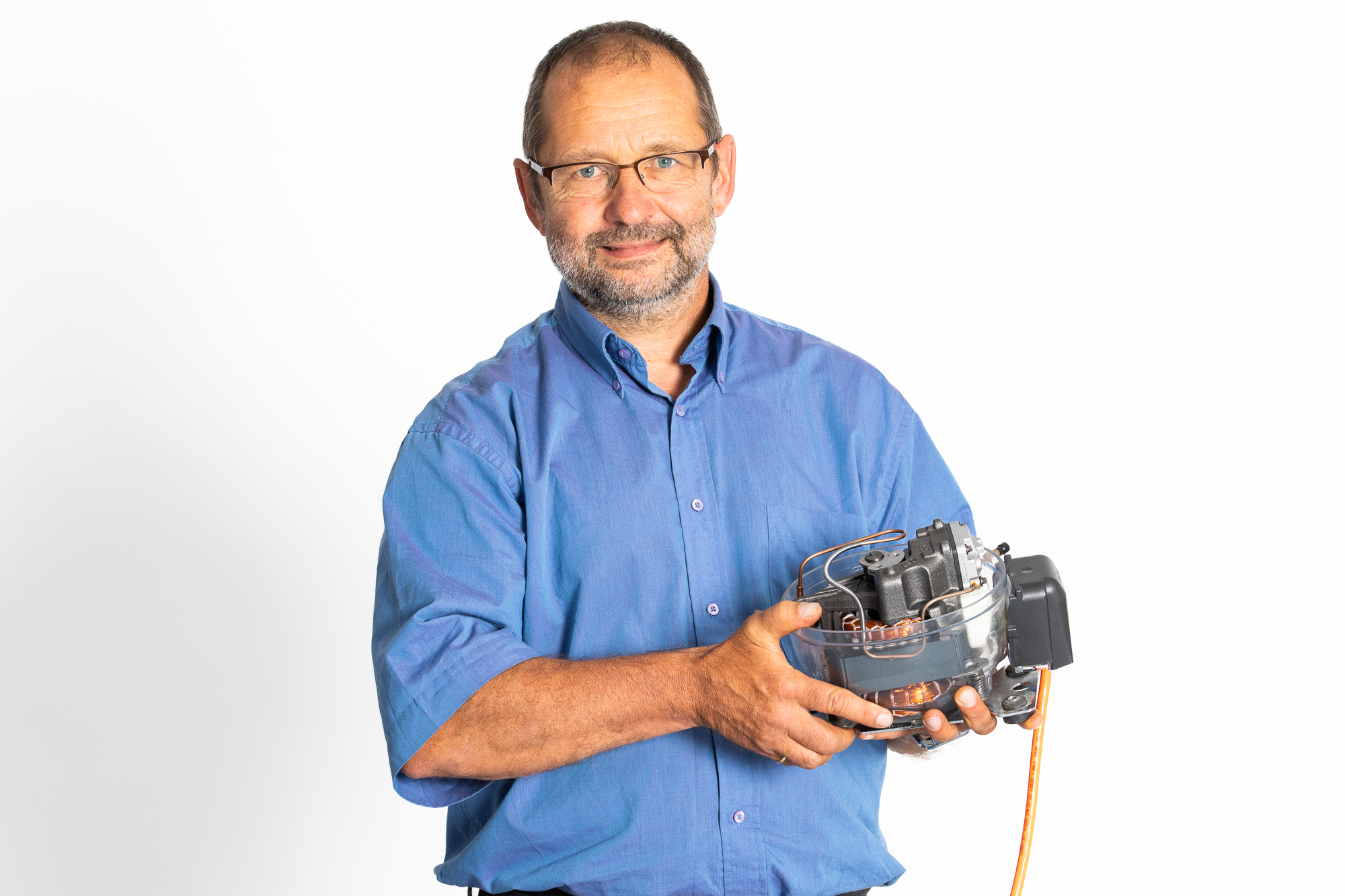 Ein Mann in einem blauen Hemd hält ein mechanisches Gerät mit Kabeln und Bauteilen in der Hand – das möglicherweise zur Messung der Heizlast oder des Energiebedarfs dient –, während er vor einem schlichten weißen Hintergrund steht. at Hochschule Coburg
