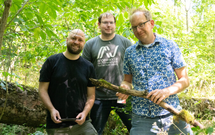 Drei Männer stehen in einem sonnendurchfluteten Wald und verkörpern den Geist von „Europas Wälder“, während sie Stöcke in den Händen halten und lächeln. Einer trägt ein schwarzes Hemd, ein anderer ein graues Hemd mit Aufdruck und der dritte ein blaues, gemustertes Hemd. Im Hintergrund sind grüne Blätter und ein umgestürzter Baum zu sehen, die ihre Verbundenheit mit der Natur und der modernen Forstpraxis widerspiegeln. at Hochschule Coburg