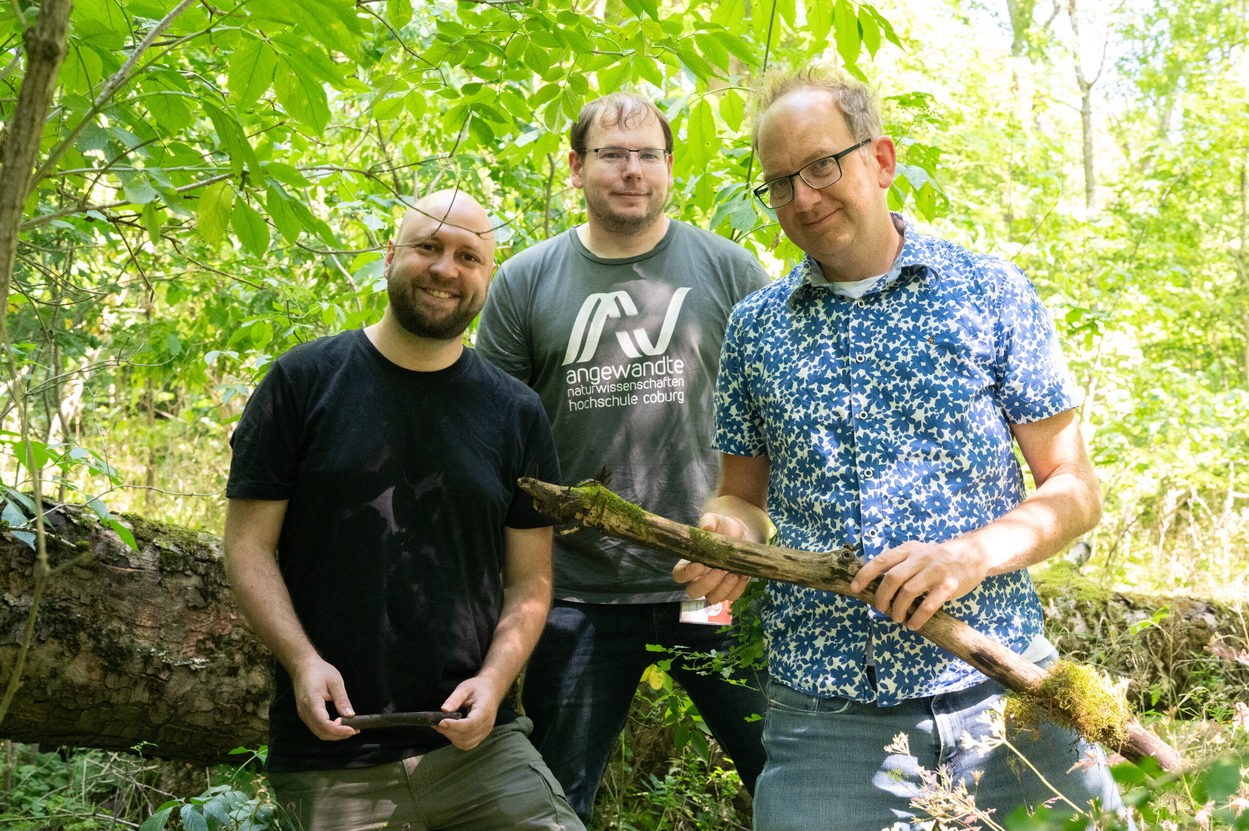 Drei Männer stehen in einem sonnendurchfluteten Wald und verkörpern den Geist von „Europas Wälder“, während sie Stöcke in den Händen halten und lächeln. Einer trägt ein schwarzes Hemd, ein anderer ein graues Hemd mit Aufdruck und der dritte ein blaues, gemustertes Hemd. Im Hintergrund sind grüne Blätter und ein umgestürzter Baum zu sehen, die ihre Verbundenheit mit der Natur und der modernen Forstpraxis widerspiegeln. at Hochschule Coburg
