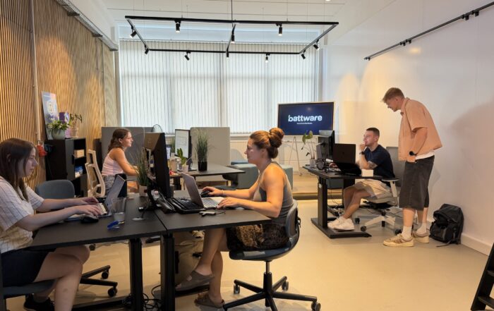 In einem hellen, modernen Büro sitzen fünf Personen an ihren Schreibtischen. Einige arbeiten an ihren Laptops, während eine Person neben einem sitzenden Kollegen steht und vielleicht über einen automatisch gespeicherten Entwurf spricht. Im Hintergrund ist auf einem Bildschirm „battware“ zu sehen. at Hochschule Coburg