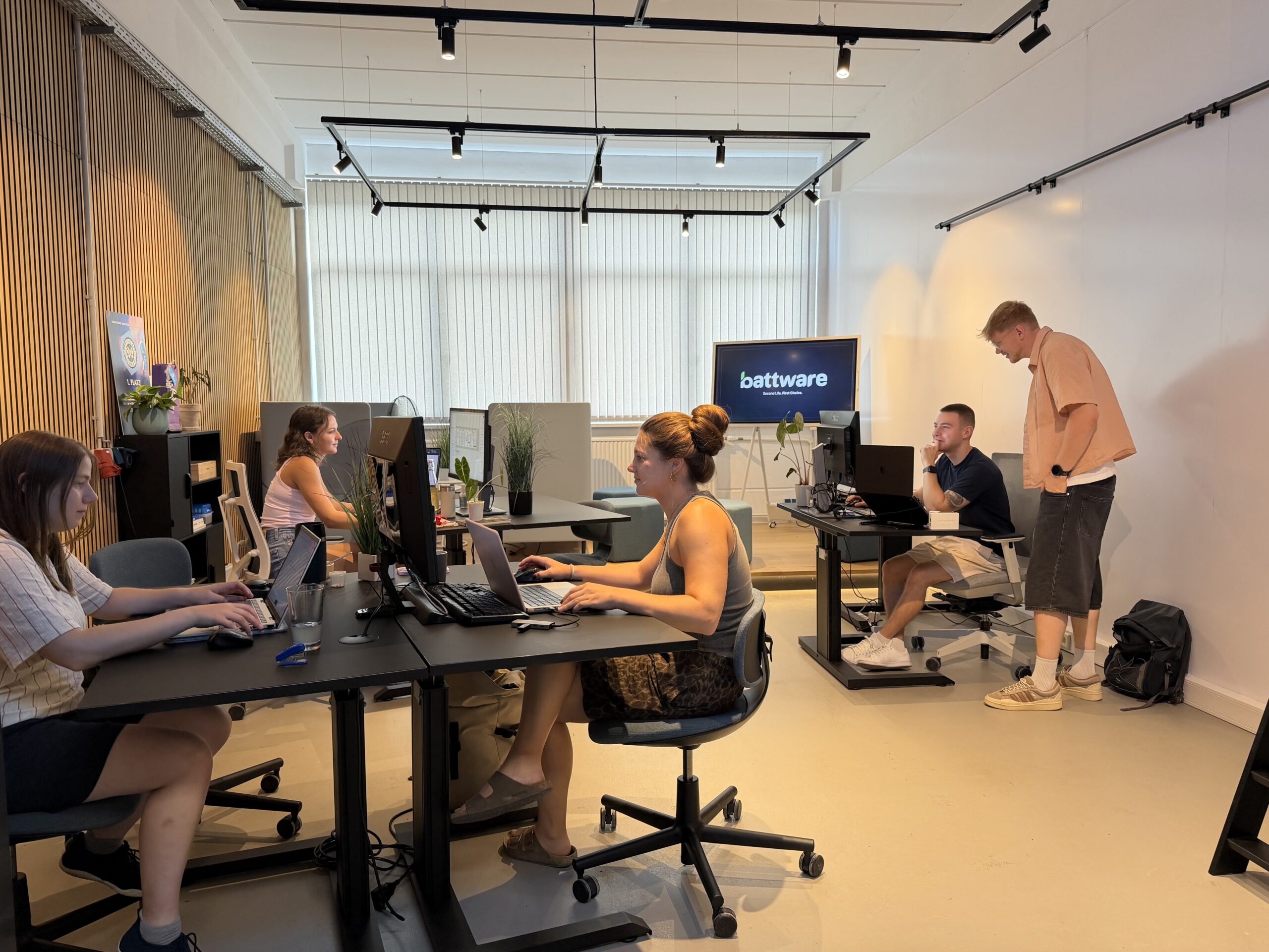 In einem hellen, modernen Büro sitzen fünf Personen an ihren Schreibtischen. Einige arbeiten an ihren Laptops, während eine Person neben einem sitzenden Kollegen steht und vielleicht über einen automatisch gespeicherten Entwurf spricht. Im Hintergrund ist auf einem Bildschirm „battware“ zu sehen. at Hochschule Coburg