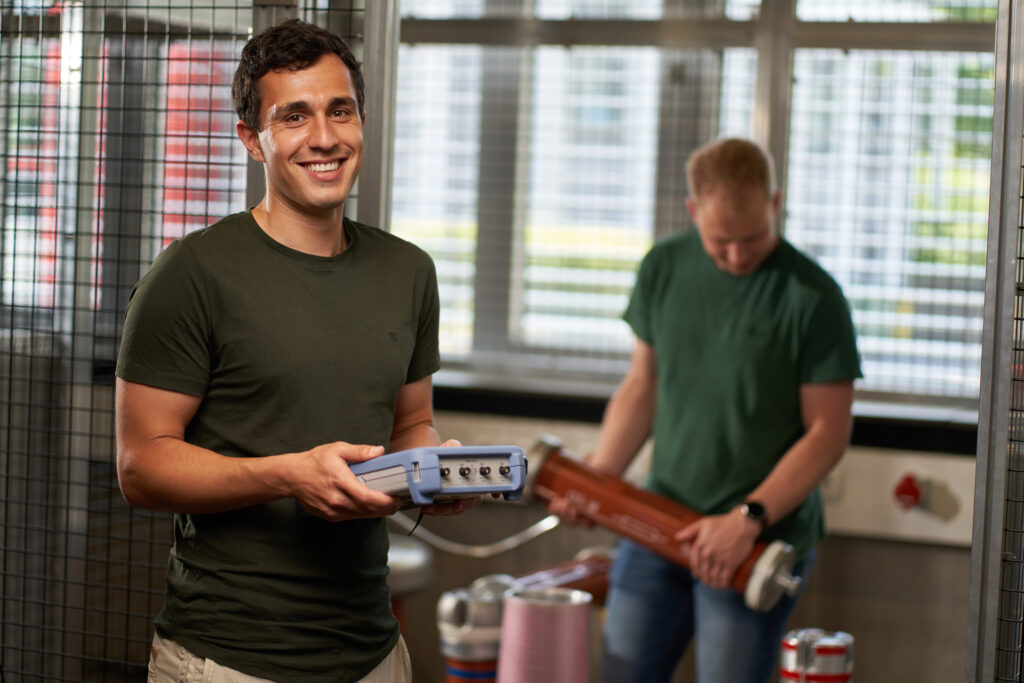 A smiling man holds electronic equipment in the foreground, while another man in the background examines a large cylindrical object in a well-lit room with metal grating and windows. at Hochschule Coburg