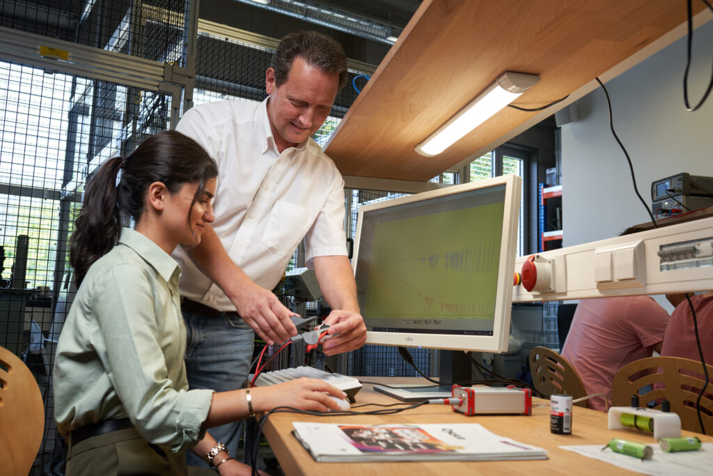 A woman sits at a desk using a computer while a man stands beside her, assisting with electronic equipment. Technical devices, cables, and documents are visible on the desk in a lab setting. at Hochschule Coburg
