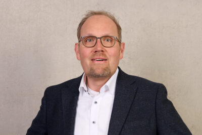 Ein Mann mit Brille, kurzem Bart und zurückgehendem Haaransatz steht vor einer schlichten beigen Wand; er trägt einen dunklen Blazer über einem weißen Hemd – seine Haltung und sein selbstbewusstes Auftreten lassen an jemanden denken, der an Aufgaben in der Zentralverwaltung gewöhnt ist –, die Hände in die Hüften gestemmt, und blickt in die Kamera. at Hochschule Coburg