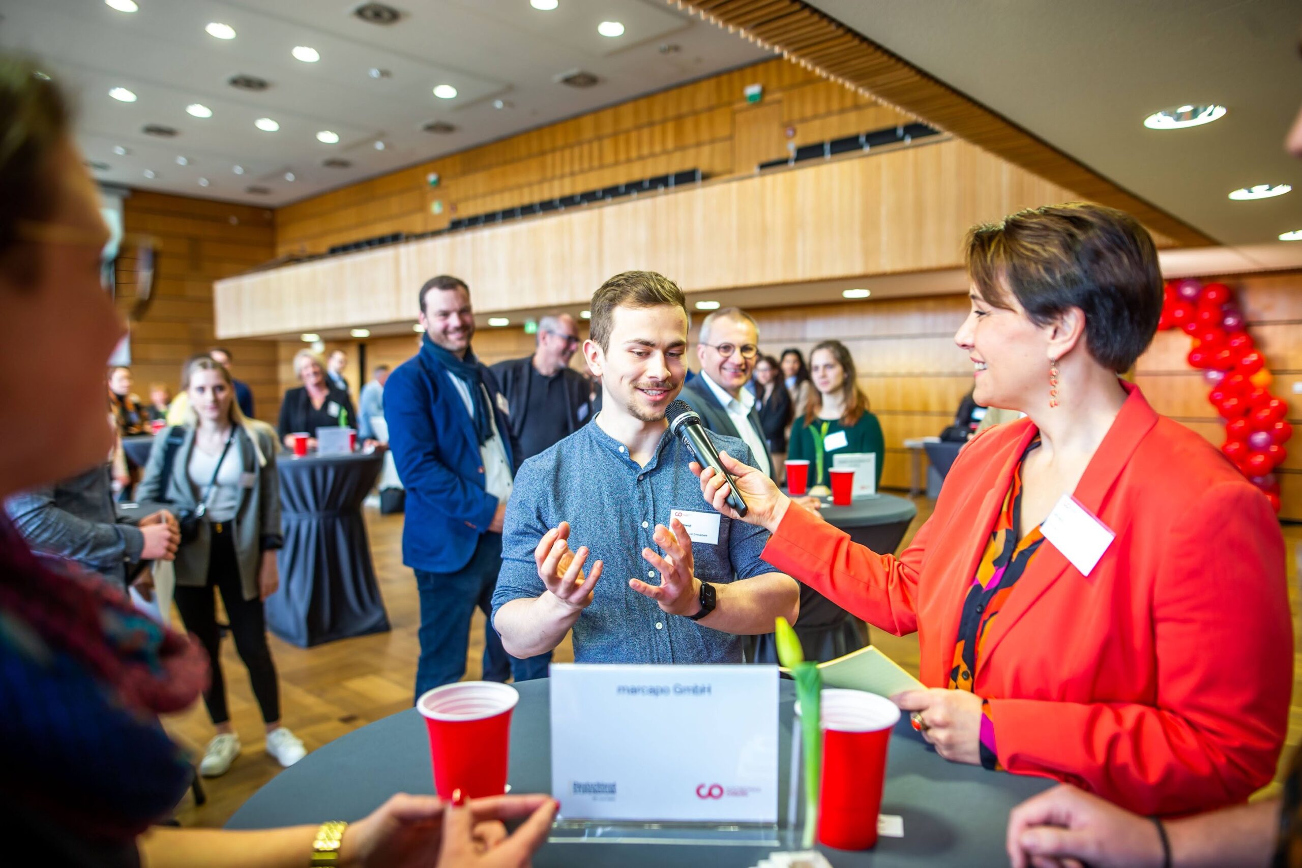 Eine Frau in einer roten Jacke interviewt einen lächelnden Mann bei einer Networking-Veranstaltung für Stipendiaten des Deutschland-Stipendiums; andere stehen um Tische mit roten Bechern herum und hören zu – in einem hellen, modernen Konferenzraum, der Potenzial fördert. at Hochschule Coburg