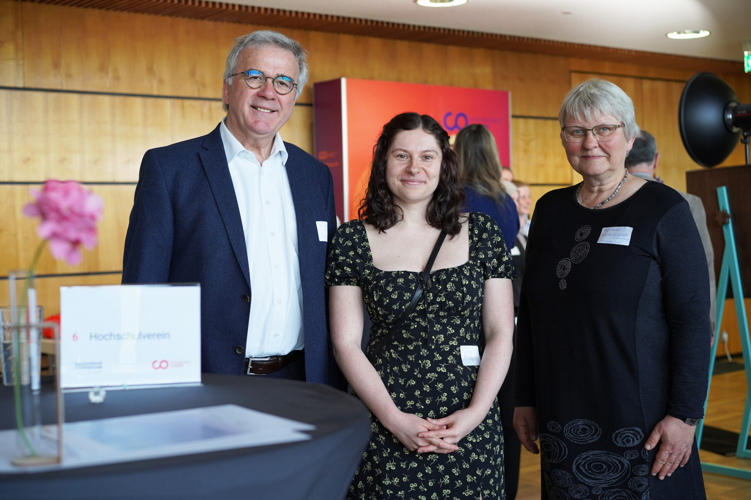 Drei Personen, darunter Studierende und ein Förderer, stehen in einem Raum in der Nähe eines Tisches mit einem Schild „Deutschland-Stipendium“ und lächeln in die Kamera. Im Hintergrund sind Holzvertäfelungen zu sehen und weitere Personen, die sich unterhalten. Die Atmosphäre wirkt formell oder professionell. at Hochschule Coburg