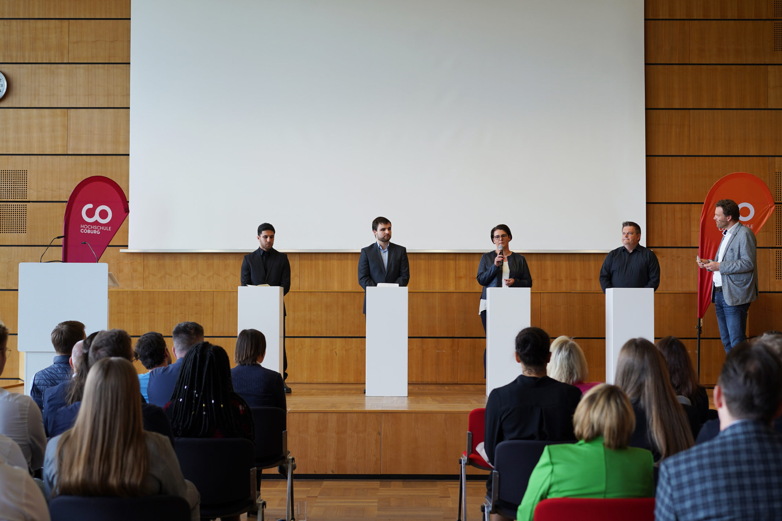 Fünf Personen stehen auf einer Bühne hinter Rednerpult und sprechen vor einem Publikum aus Studierenden und Förderern in einem modernen Konferenzraum. Im Hintergrund sind zwei rote Banner und eine große leere Leinwand zu sehen. Das Publikum, darunter auch Empfänger des Deutschland-Stipendiums, sitzt und hört aufmerksam zu. at Hochschule Coburg