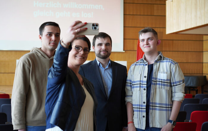 Vier Personen posieren für ein Selfie in einem Raum mit Holzwänden und roten Stühlen; auf einem Bildschirm im Hintergrund ist der deutsche Text „Herzlich willkommen, gleich geht es los!“ zu sehen – ein lebhafter Moment unter Studierenden und Förderern bei einer Veranstaltung des Deutschland-Stipendiums. at Hochschule Coburg