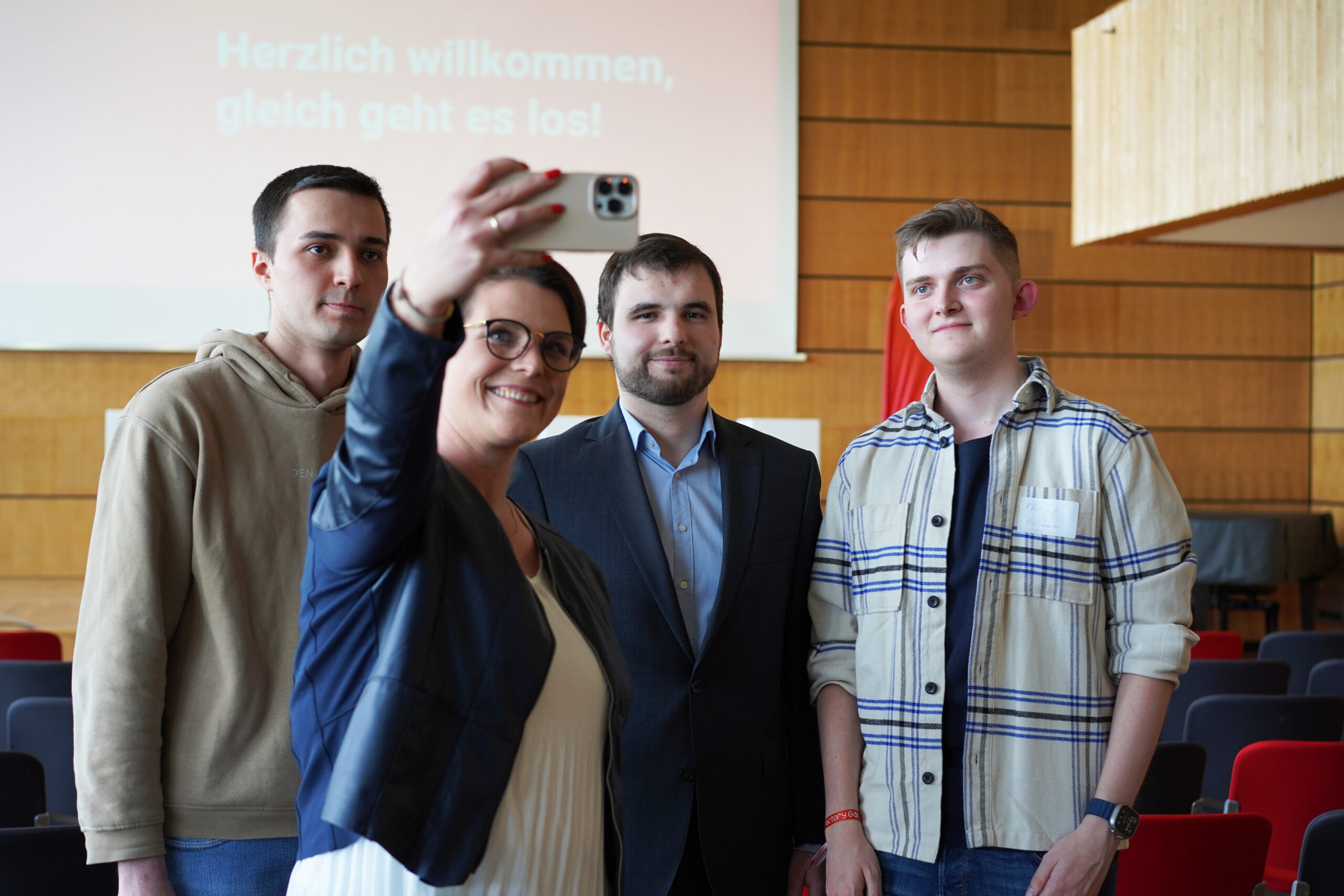 Vier Personen posieren für ein Selfie in einem Raum mit Holzwänden und roten Stühlen; auf einem Bildschirm im Hintergrund ist der deutsche Text „Herzlich willkommen, gleich geht es los!“ zu sehen – ein lebhafter Moment unter Studierenden und Förderern bei einer Veranstaltung des Deutschland-Stipendiums. at Hochschule Coburg