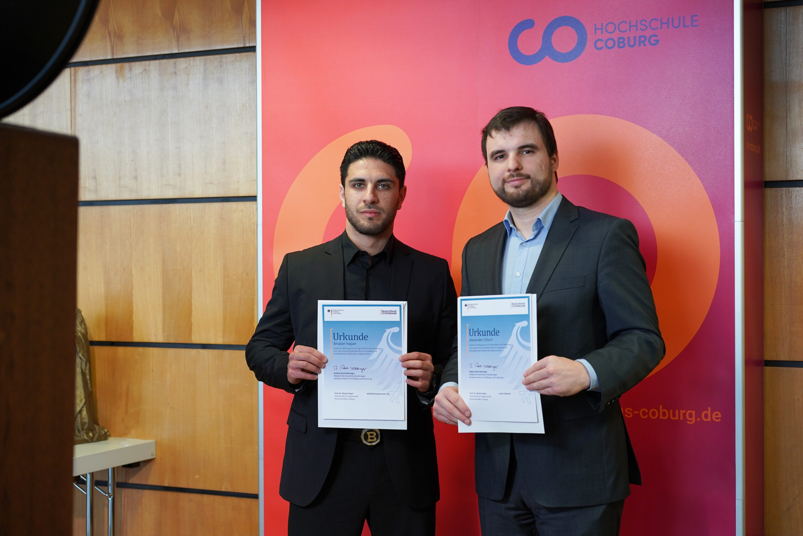 Zwei Männer in Anzügen halten Urkunden in den Händen, während sie vor einem Banner der Hochschule Coburg stehen und ihre Auszeichnung als Förderer des Deutschland-Stipendiums feiern. Sie posieren für ein Foto, blicken in die Kamera und haben im Hintergrund eine Holzwand sowie ein rotes Display im Hintergrund. at Hochschule Coburg