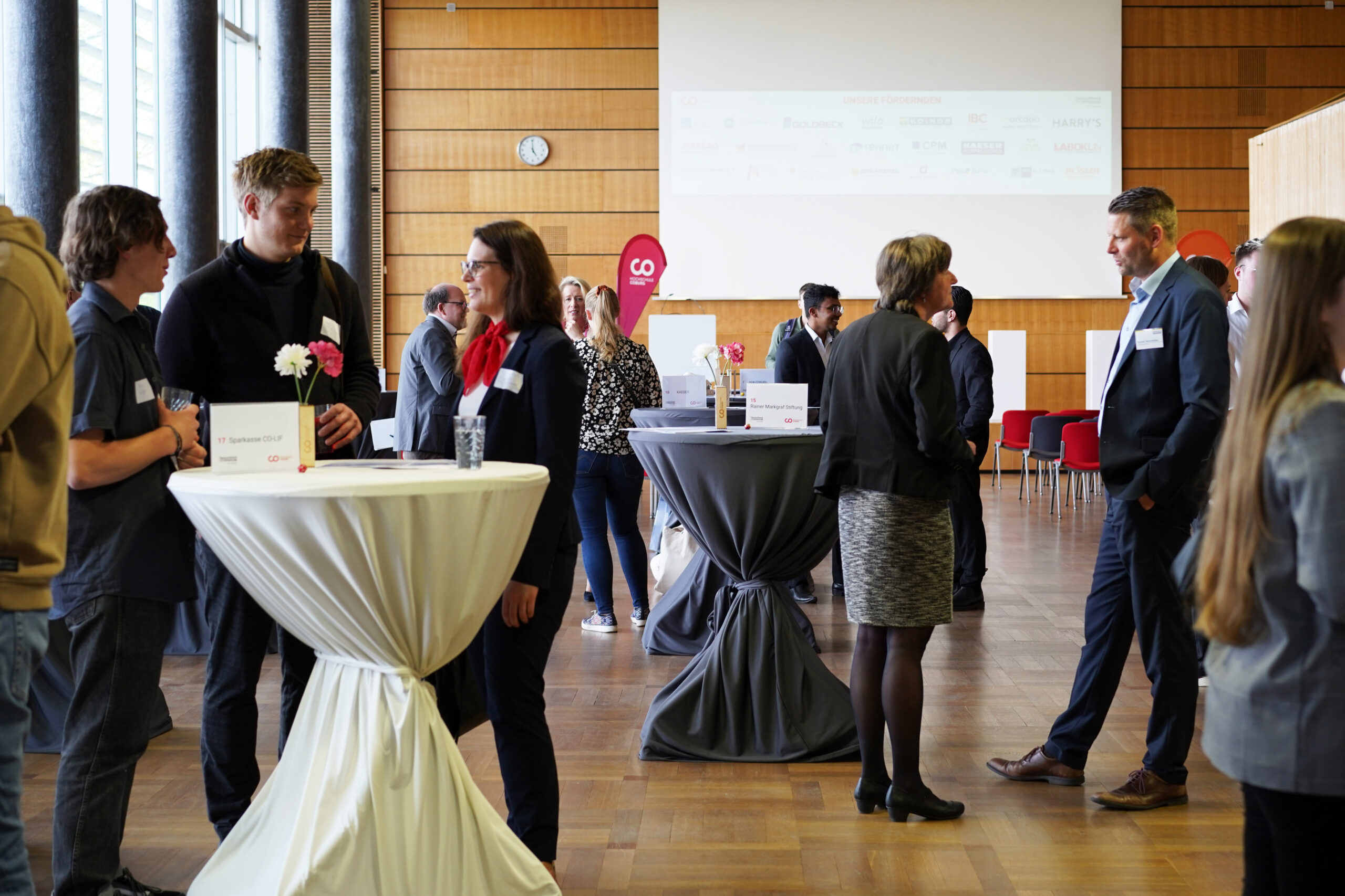 In einem geräumigen, modernen Raum mit Holzwänden, großen Fenstern und einer Präsentationsleinwand stehen Menschen in Geschäftskleidung um Cocktailtische herum und unterhalten sich. Auf den Tischen liegen Namensschilder und Hinweisschilder zum Deutschland-Stipendium, was deutlich macht, dass es sich um eine Veranstaltung handelt, bei der Studierende und Förderer miteinander in Kontakt treten und sich vernetzen. at Hochschule Coburg